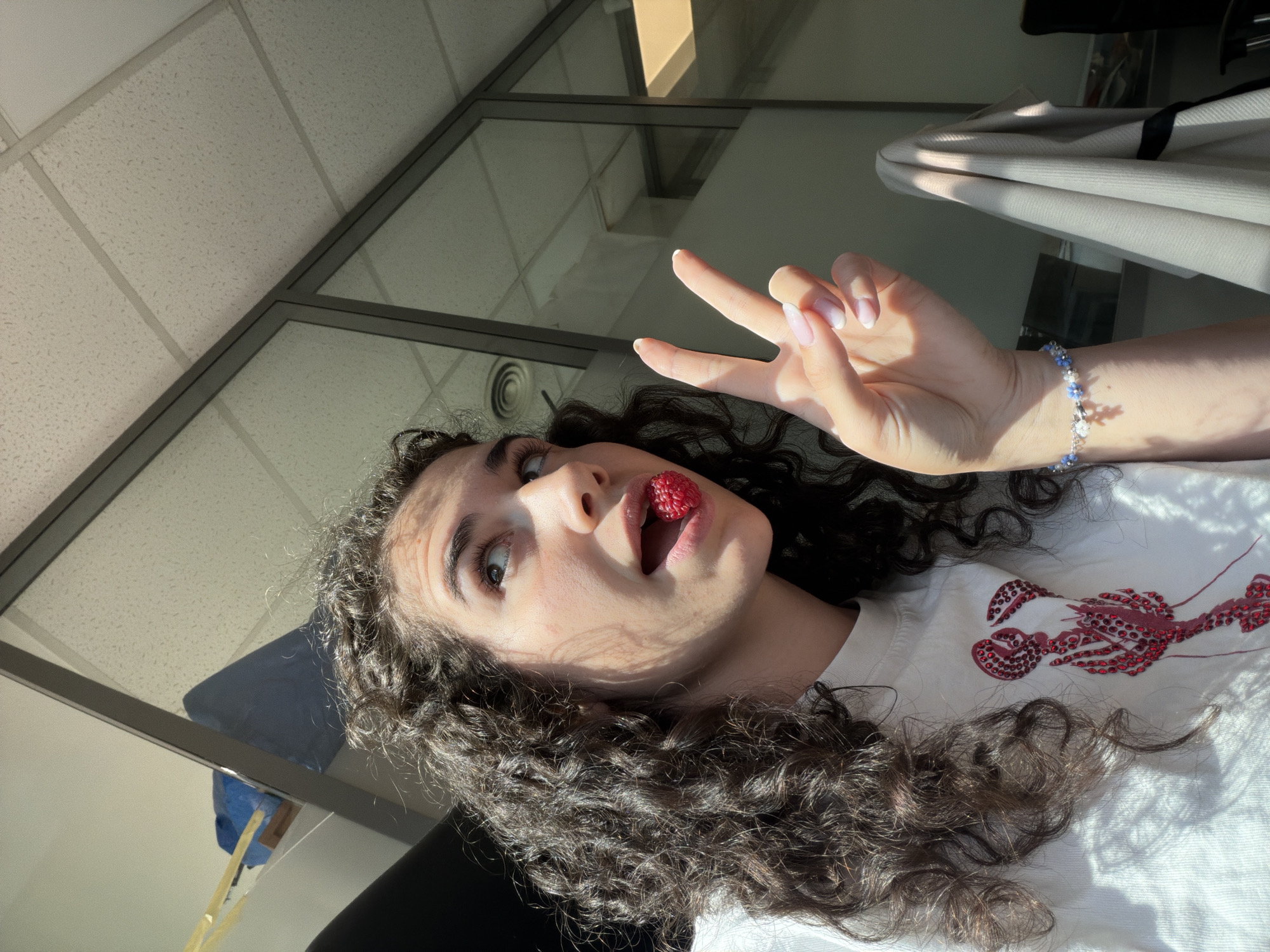 In an indoor office environment, a young woman with curly dark hair is captured from the chest up. She is looking upwards and to her right, with a raspberry perched on her tongue and her mouth slightly open. Her right hand is raised, forming a peace sign. She is wearing a white t-shirt adorned with a red, beaded lobster design on the left side of her chest. A bracelet with blue and white beads is visible on her wrist. The background consists of a ceiling with fluorescent lighting and panels, and a glass partition with reflections of the outside, suggesting a modern office space. Sunlight casts shadows and highlights across the scene. The location in the city of Sofia, Bulgaria, is not discernible from the image itself.