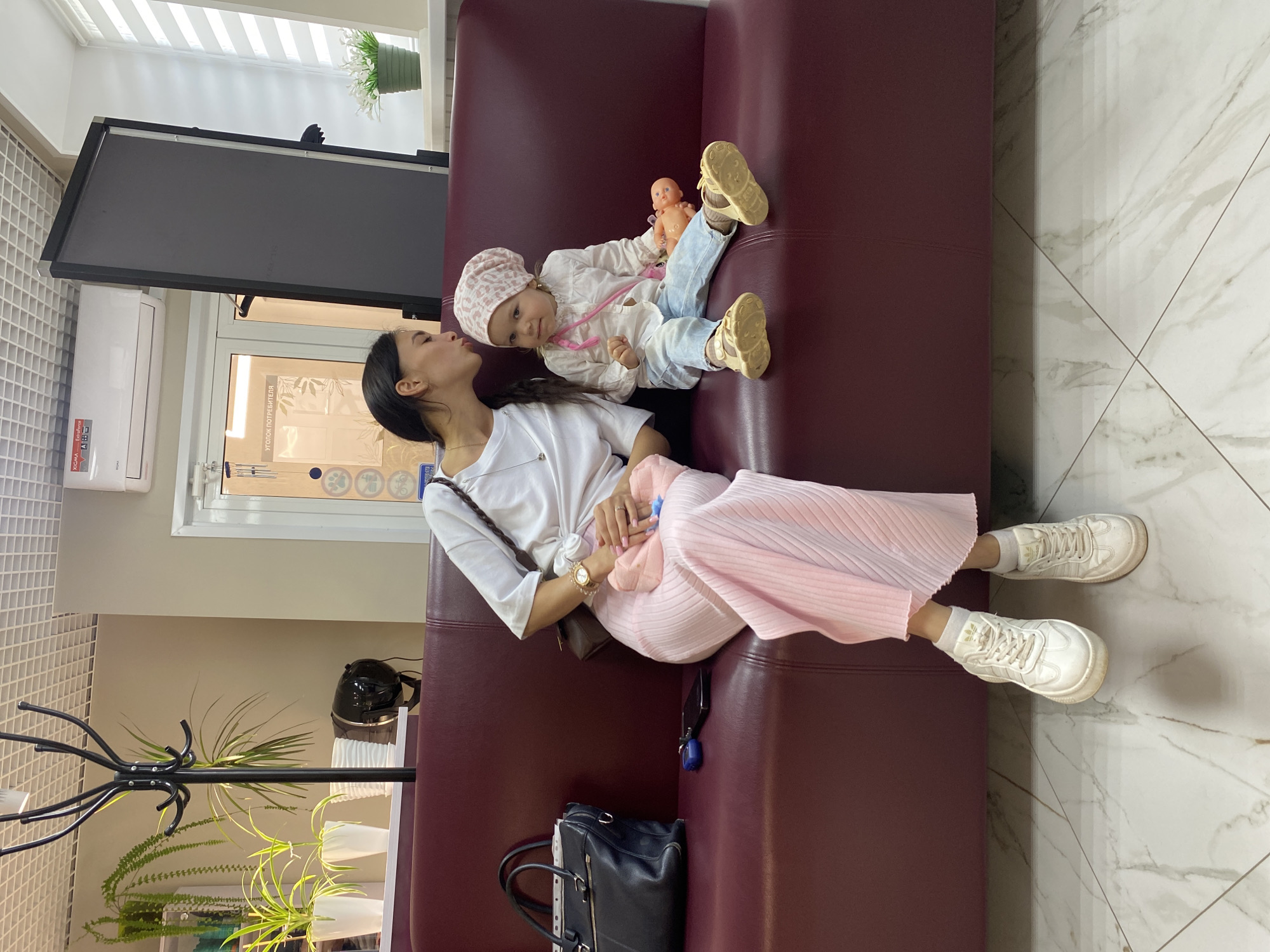 The image shows a young woman sitting on a burgundy couch, with a young child sitting next to her. The woman is wearing a white t-shirt and a pink pleated skirt, and she is leaning down to kiss the child on the forehead. The child is wearing a light-colored shirt, blue jeans, and a pink headscarf. The child is holding a small doll. In the background, there is a black coat rack with plants, a black handbag on the couch, a smartphone, and a blue keychain. The room appears to be a waiting area, possibly in a medical facility, with a tiled floor and a window with blinds. The location is identified as Chaikovsky, Russia.