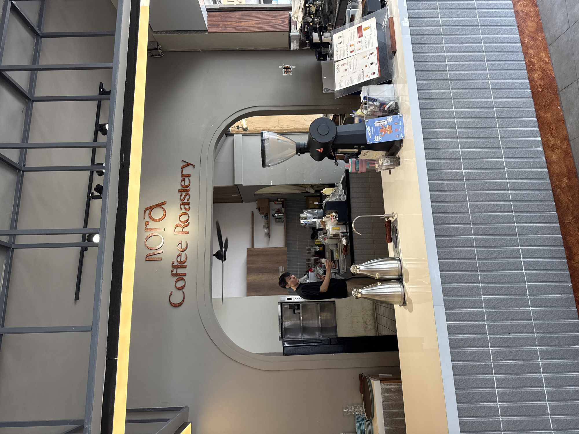 The image captures an interior view of a coffee roastery in Nha Trang Ward, Vietnam. In the background, behind an arched entryway, a male barista in a black t-shirt and beige pants is preparing drinks at a counter. Shelving and kitchen equipment are visible. A large coffee grinder stands prominently on the foreground counter, next to a menu display. The counter itself is topped with a light-colored surface and features two silver pour-over dispensers and a faucet. The facade of the counter is covered in a textured gray tile pattern. The roastery's name, "nora Coffee Roastery," is displayed in large, reddish-orange lettering above the arched entrance. The overall lighting is bright and artificial, suggesting an indoor commercial setting.