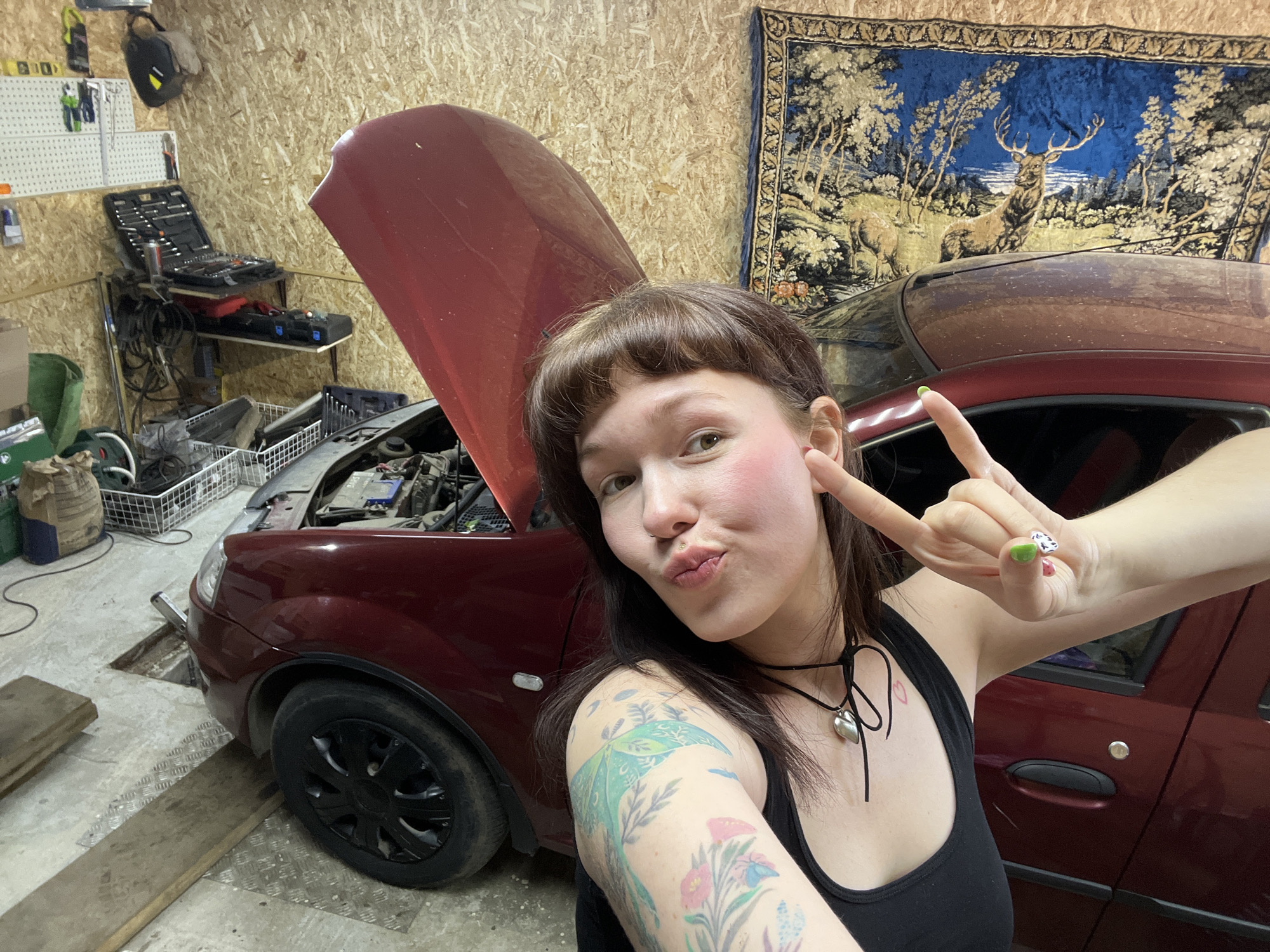 A young woman poses for a selfie in what appears to be a garage. She is positioned in front of a burgundy car with its hood open. She is wearing a black tank top and has a black choker around her neck. Her left arm, visible in the foreground, is tattooed with colorful floral and insect designs. Her hair is brown with bangs. She is making a puckered-lip expression and a "rock on" hand gesture with her right hand.
Behind her, a tapestry depicting a scene with deer hangs on a wood-paneled wall. A shelving unit with tools and equipment is visible in the background to the left. The garage floor appears to be concrete, and the car is partially elevated on wooden planks.
There is no evidence to confirm that the location of the image is in the city of Chaikovsky, Russia.
