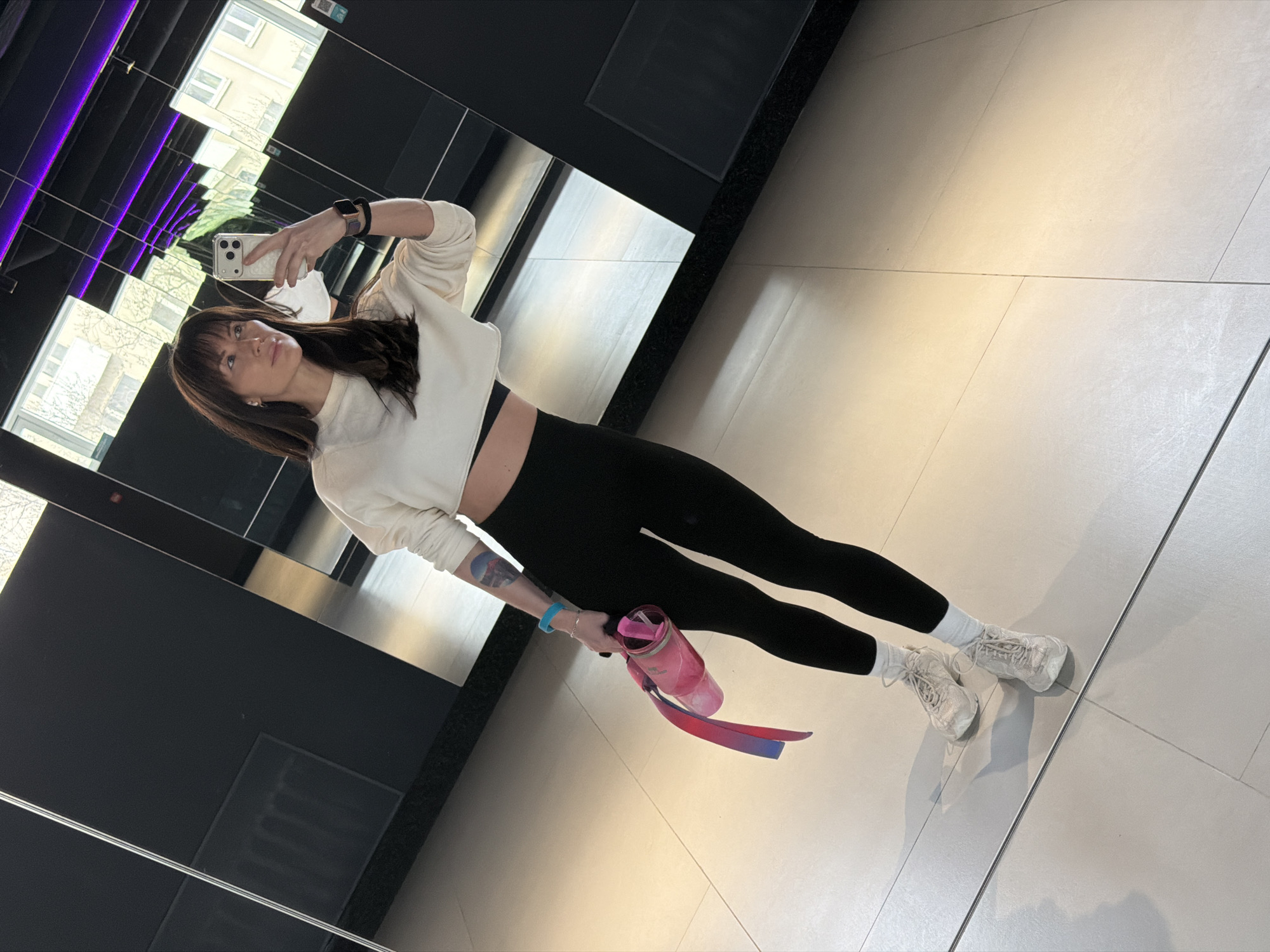 A woman is taking a selfie in a mirror. She is wearing a white cropped sweater, black leggings, white socks, and beige sneakers. She is holding a water bottle in her left hand. The location appears to be a modern-looking building with black and silver reflective surfaces. The reflection shows buildings outside. The location may be in Perm, Russia.