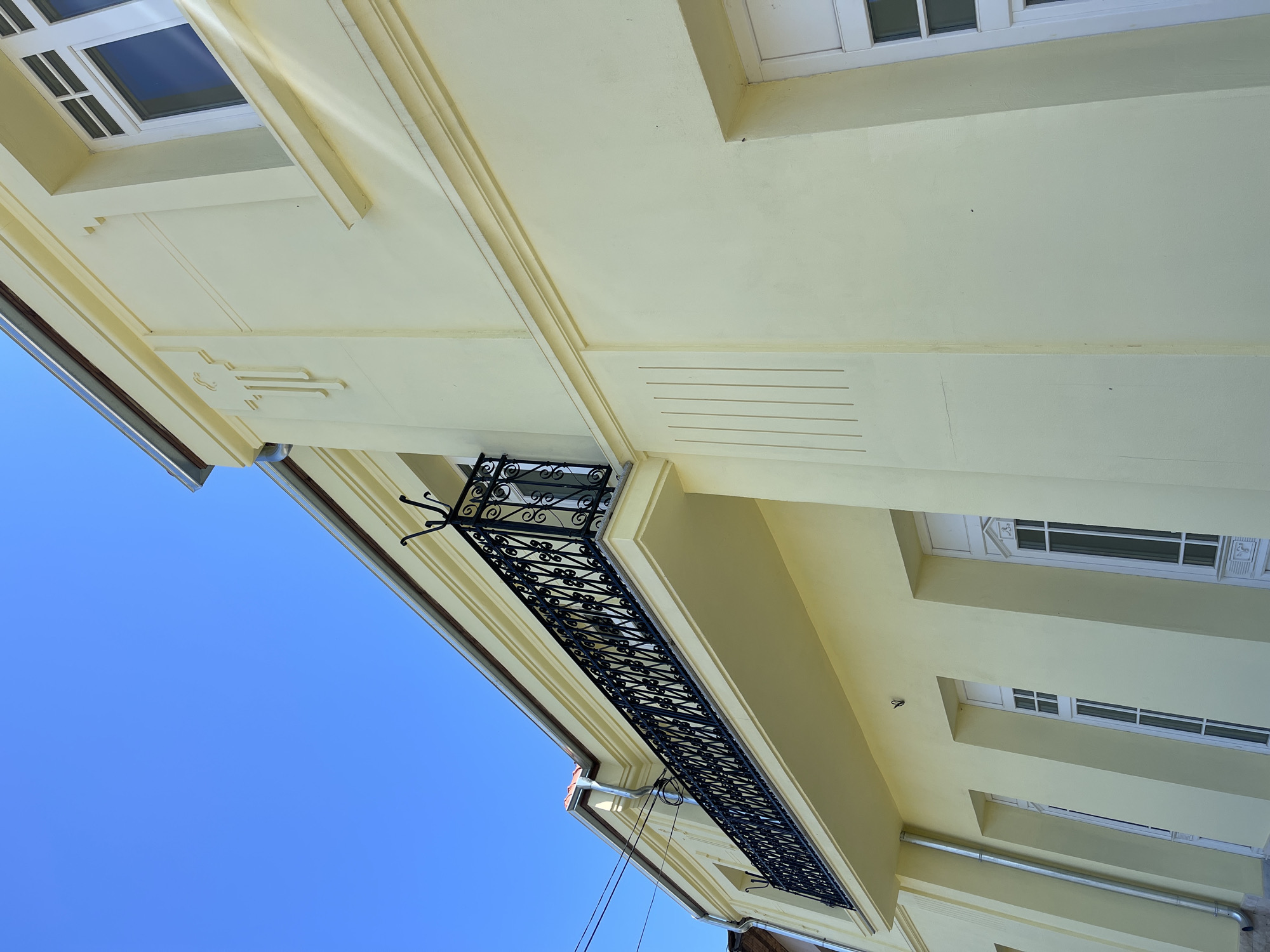 The image depicts a low-angle view of a pale yellow building in Somovit, Bulgaria. The building's architecture features decorative trim, including recessed lines and geometric accents. Windows with white frames are visible. A black wrought-iron balcony is prominent. The sky is blue. Electrical wires are visible.