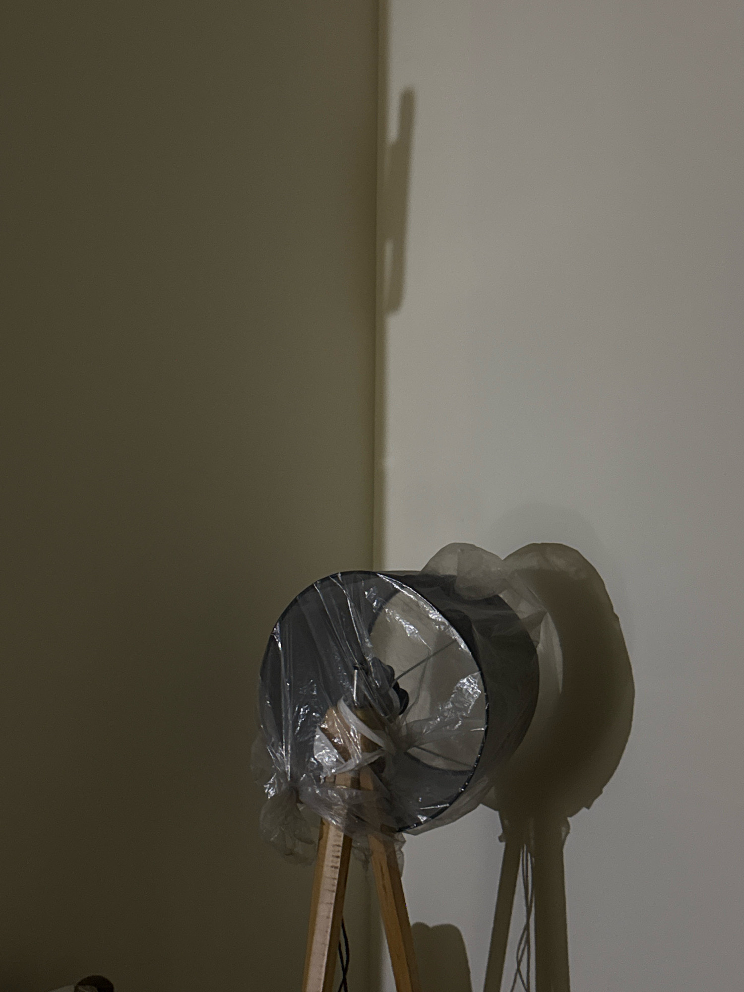 A floor lamp, covered in plastic, stands near a wall in a dimly lit room. The lamp has a wooden tripod base and a dark lampshade. A shadow of the lamp and a part of the wall is cast on the adjacent wall. The scene appears to be indoors, possibly in a residential setting. There are no people visible in the image. The location of Kohistan Enclave, Pakistan is not ascertainable from the image.
