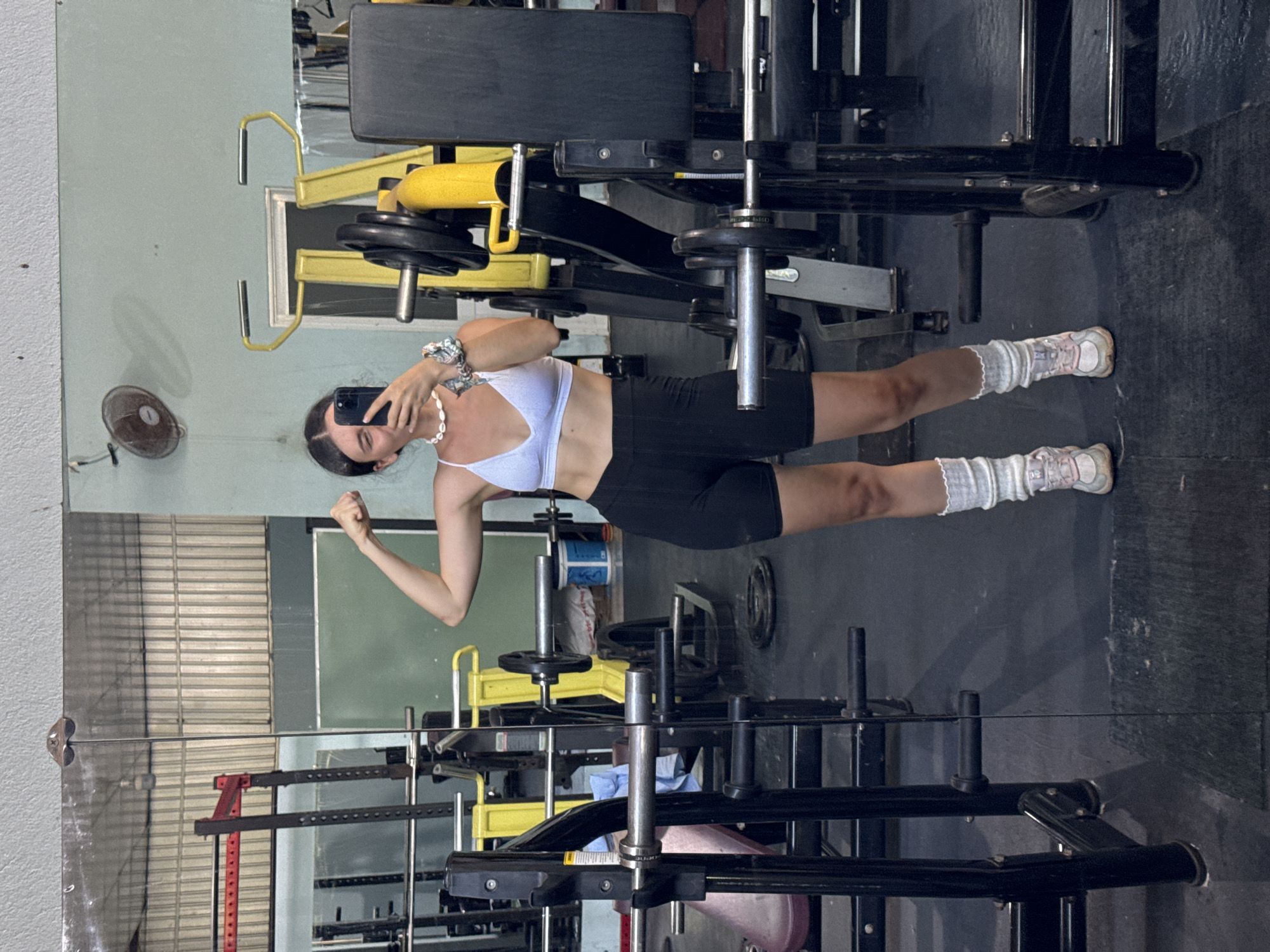 A young woman is reflected in a mirror in a gym in Mũi Né, Vietnam. She is holding a smartphone and appears to be taking a selfie. She is wearing a white sports bra, black shorts, scrunched socks, and athletic shoes. The gym has exercise equipment, including weight machines with yellow accents. The floor is dark, and there is a fan on the wall. The reflection suggests the gym has large mirrored surfaces and is moderately lit.