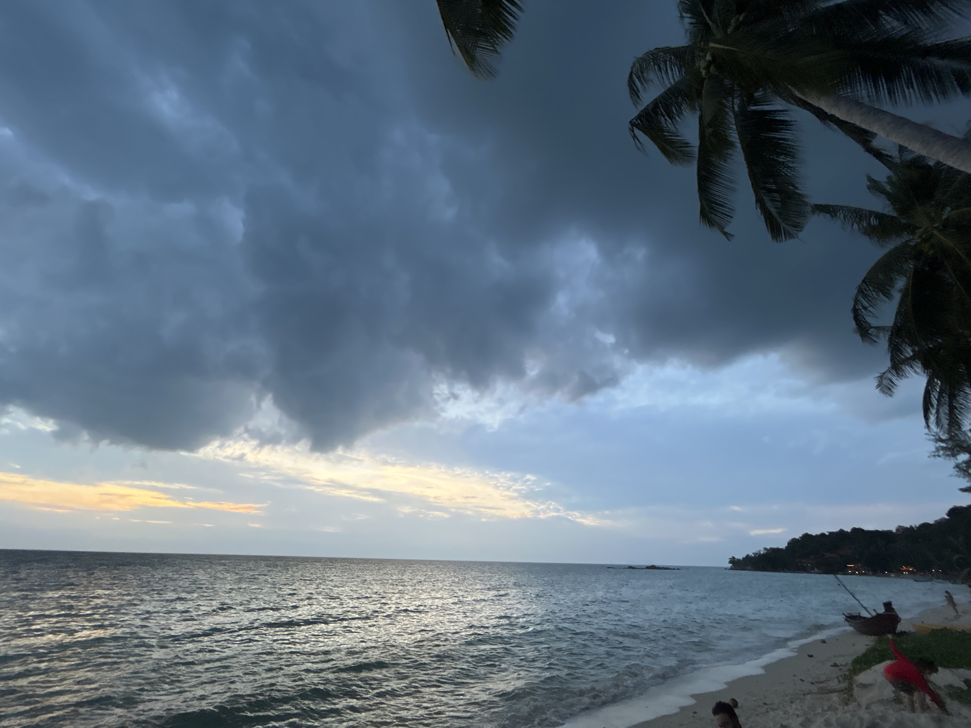 A wide-angle, low-angle shot captures a beach on Ko Pha-ngan District, Thailand, under a dramatic, cloud-filled sky. The ocean, a deep, textured blue, stretches to the horizon where a faint glow of sunset peeks through the heavy clouds. Palm fronds from two trees frame the top right and left corners of the image, their dark silhouettes contrasting with the lighter sky. The sandy shore in the foreground is dotted with a few scattered objects and people. A lone, dark wooden boat rests on the sand, its fishing net draped over the side. Further down the beach, a small figure in red appears to be running or playing. Another figure is partially visible in the lower left corner, seated on the sand. The water laps gently at the shore, creating small, foamy waves. The overall atmosphere is moody and serene, with the encroaching darkness of the storm clouds suggesting an imminent change in weather.