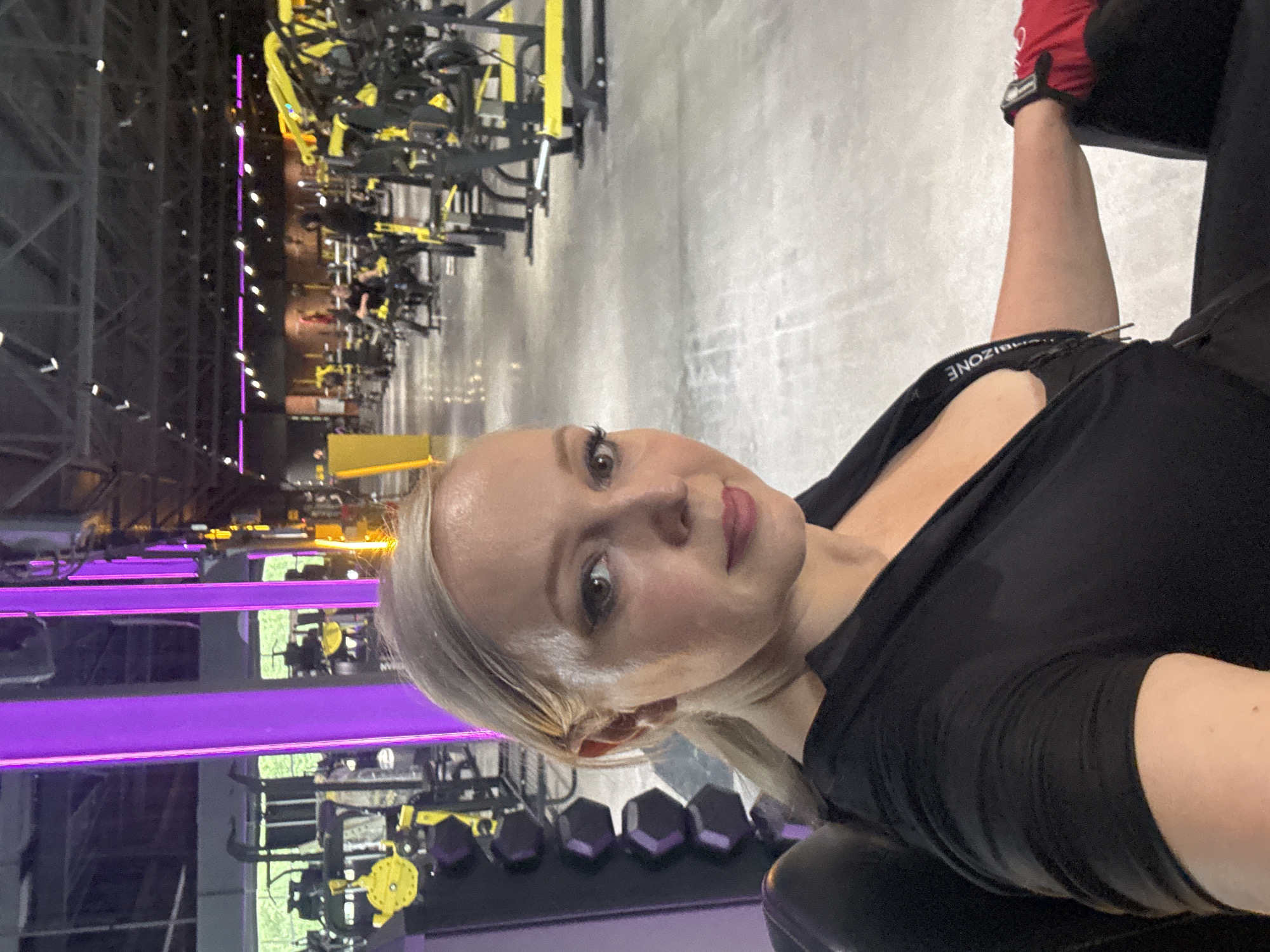 The image shows a woman with blonde hair pulled back in a ponytail, wearing a black v-neck top, in a gym. She has makeup on, including eyeliner and lipstick. To her left, there is a rack of hexagonal dumbbells. The gym has a concrete floor and rows of yellow and black exercise machines. In the background, there are purple and yellow lights. The image does not provide any information about the city of Komsomolsk-on-Amur, Russia.