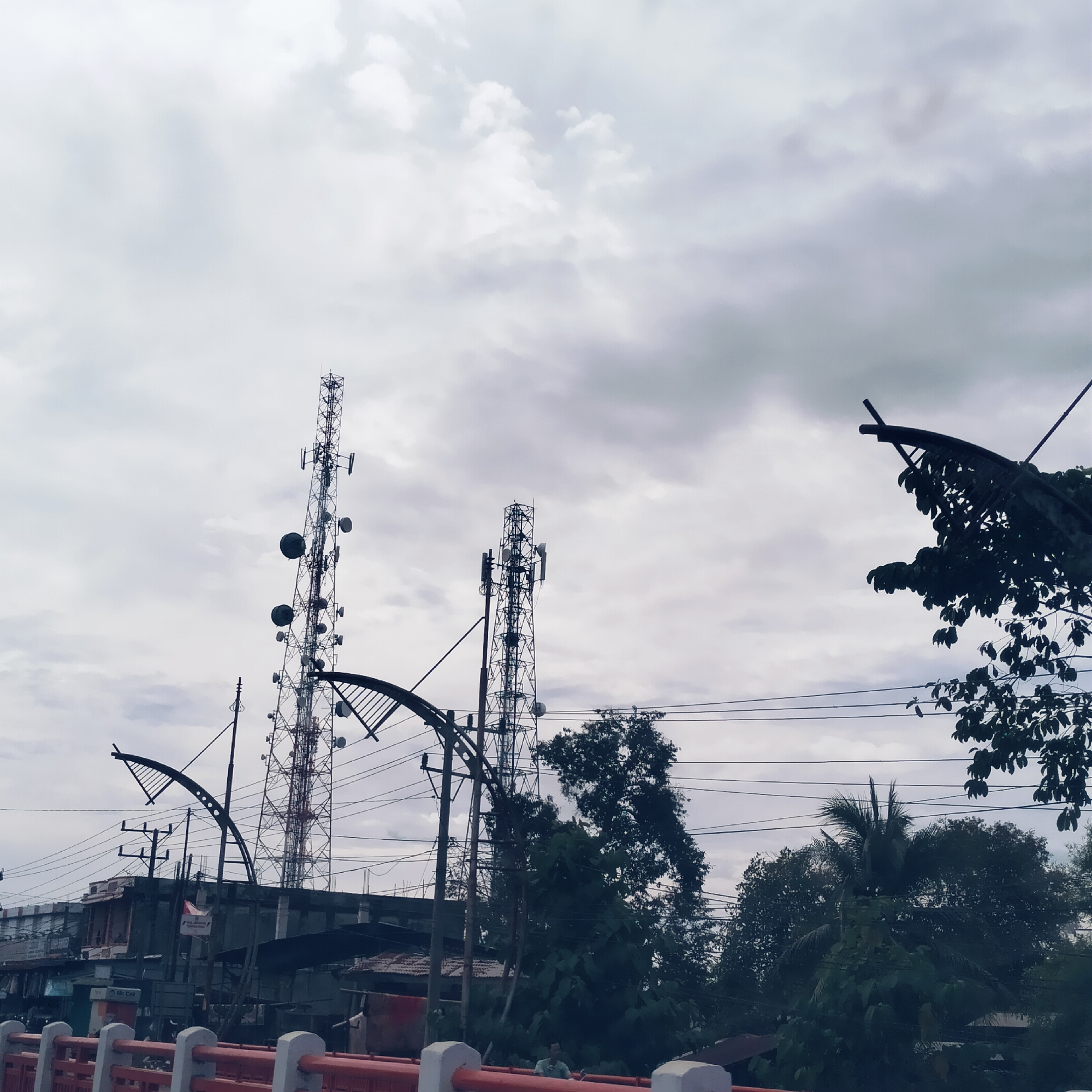 The image depicts a dense urban or semi-urban landscape under an overcast sky, likely during daytime in Baro, Indonesia. In the immediate foreground, a red and white railing suggests a bridge or elevated viewpoint. Below this railing, a person in a light-colored shirt is faintly visible.
The midground is dominated by two towering communication or cellular towers, heavily equipped with multiple antennas and large parabolic dishes. A complex network of overhead power lines and cables crisscrosses the scene, supported by utility poles and intertwined with streetlights featuring decorative, arched designs.
Beneath the intricate web of wires and towers, various low-rise buildings and structures are visible, some appearing functional or commercial. On one of these structures, a white sign with the text "Mc Cell" is discernible. Lush green trees and dense foliage are interspersed throughout the environment, partially obscuring some buildings and adding a natural element to the built infrastructure. The sky is largely covered by grey, heavy clouds, contributing to the subdued lighting of the scene. The overall impression is one of robust, yet sprawling, telecommunications and power infrastructure in a populated area.