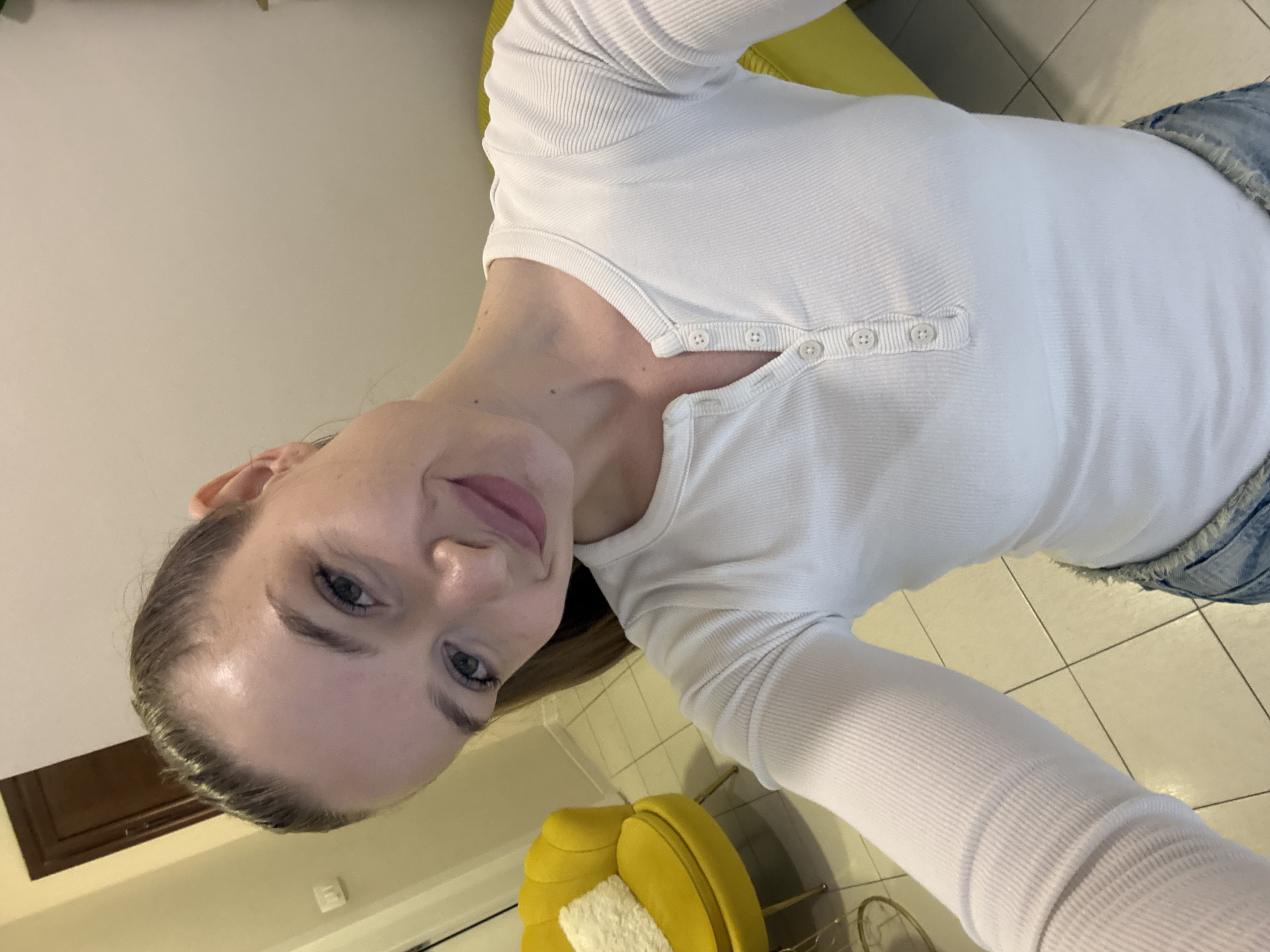 A woman with fair skin and light brown hair pulled back is seated on a bright yellow chair with a white cushion. She is wearing a white, long-sleeved shirt with button details. Tile flooring is visible. The location could potentially be a residential interior in Budva, Montenegro.