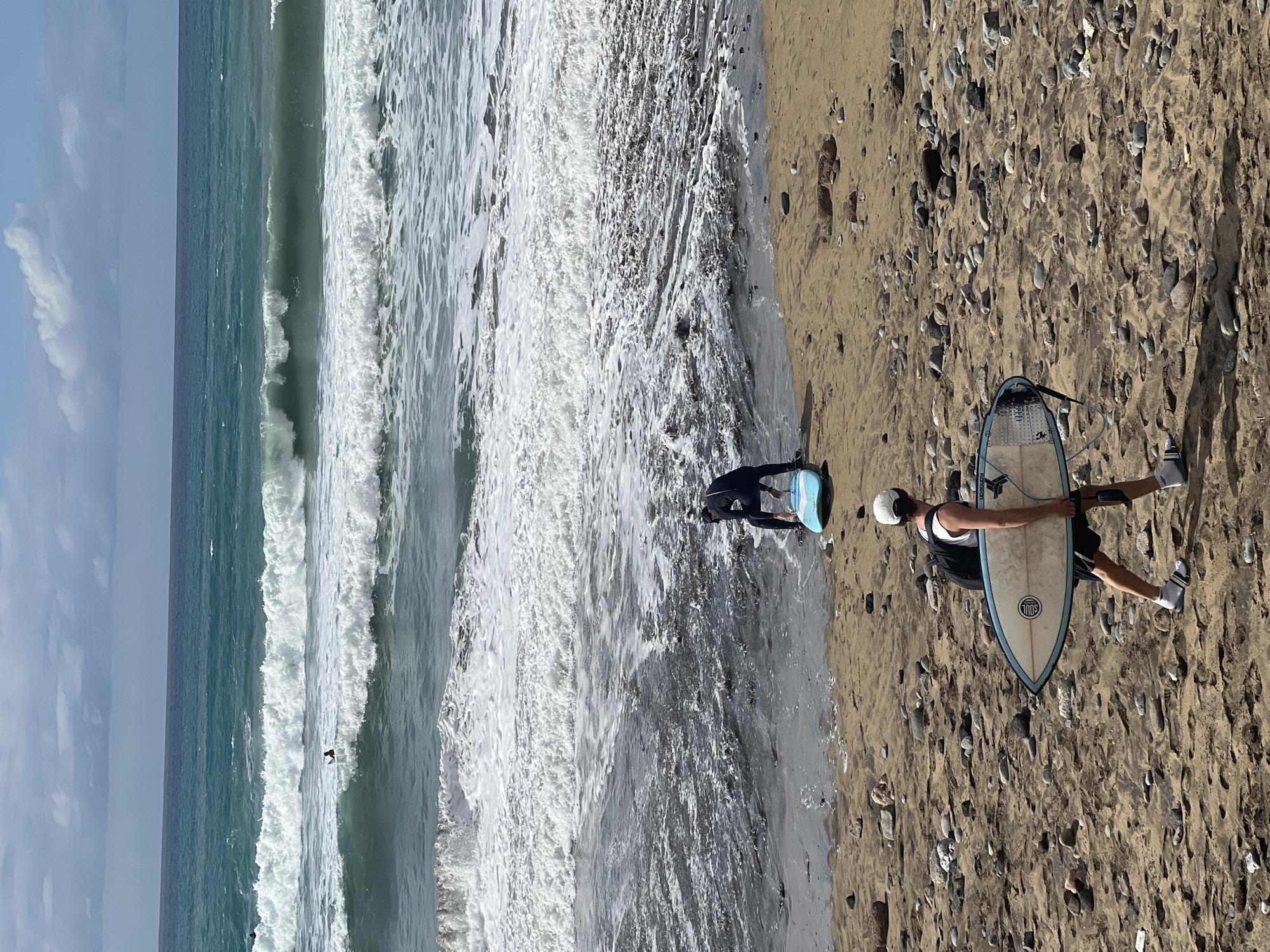 This image captures a vibrant beach scene, likely in Las Palmas, Spain, presented in a vertical orientation where the horizon runs from top to bottom.
On the leftmost side of the image, a clear, light blue sky with scattered white clouds is visible. To its right, the ocean spans the central portion, displaying distinct lines of breaking waves. The water transitions from a calmer, deeper blue-green further left to foamy white where the waves crest and break towards the right, meeting the shore. A small, dark figure appears to be in the water further out, potentially another surfer.
The rightmost section of the image features a sandy beach, densely textured with numerous small, smooth pebbles and rocks of varying shades of grey, brown, and white.
Two individuals are present, engaged in water sports activities. Closer to the bottom-right corner, a person stands on the sandy shore, holding a surfboard with both hands. This surfboard has a light-colored deck with light blue rails and features a circular logo containing the word "SOUL," along with fainter text that appears to be "WOODSON" or similar near the nose. The person is dressed in dark shorts, a light-colored top, a white cap, and white socks worn with dark sandals or slides, suggesting they are either preparing to enter or have just exited the water. Further to the left on the beach, closer to the foamy waterline, another person, clad in what appears to be a dark wetsuit or rashguard, is bending over a small, light-blue board, possibly a bodyboard or skimboard, interacting with the shallow waves.
The scene depicts bright daylight and clear, sunny weather, indicating an enjoyable day for beach activities. The overall mood conveyed is one of leisure and active engagement with the ocean.