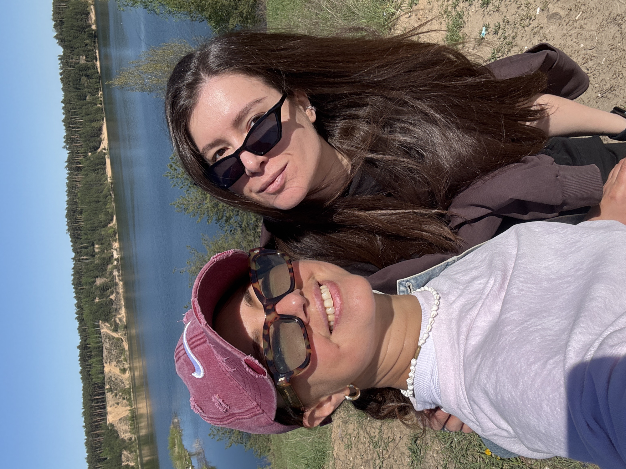 Two women are lying on the ground next to a body of water. One woman is wearing sunglasses and a brown sweatshirt. The other is wearing sunglasses, a maroon cap, and a white sweatshirt with a pearl necklace. The background includes a body of water with a forested shoreline and a blue sky. The location is likely near the city of Kazan, Russia, based on the landscape and vegetation.