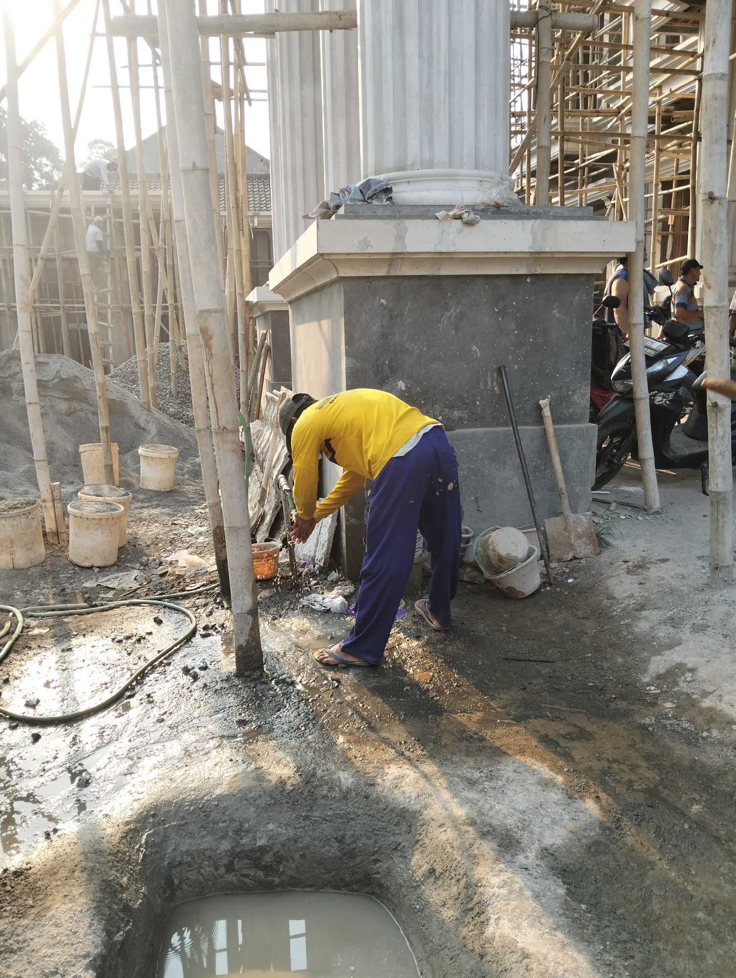 The image depicts an outdoor construction site in Durenombo, Indonesia, under bright sunlight. A person wearing a yellow long-sleeved shirt and blue pants is bent over, washing their hands under a running hose connected to a water source. The ground is muddy and wet, with a large pool of water in a dug-out area in the foreground. Several empty buckets are scattered around, along with a shovel and other construction debris. In the background, scaffolding made of bamboo is visible, suggesting ongoing construction. A motorcycle is parked near a concrete pillar. The scene conveys a sense of ongoing work and the raw environment of a construction project.