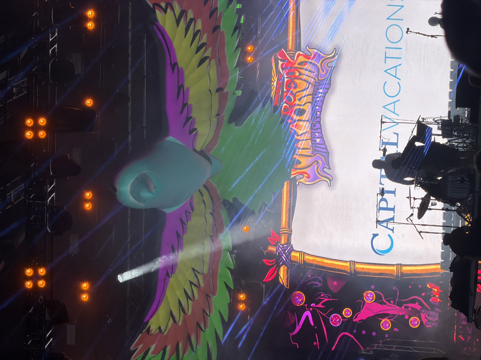 The image captures a concert stage at an outdoor event in Virginia Beach, Virginia, USA. A large, vibrant screen displays a colorful parrot graphic with wings spread wide, accompanied by a logo that appears to read "SUNBEAM MUSIC FESTIVAL" in a psychedelic font. Below the logo, the text "CAPITAL VACATIONS" is visible on the screen in light blue lettering.
In the foreground, several musicians are on stage performing. Silhouetted figures can be seen operating musical equipment, including a drummer with a set of drums and a keyboard player. Microphones and stage lighting are also prominent. The stage is further illuminated by numerous orange lights positioned above and behind the performers, creating a dramatic effect. Blue laser beams crisscross the scene, adding to the festive atmosphere. The overall impression is that of a lively and energetic music festival.