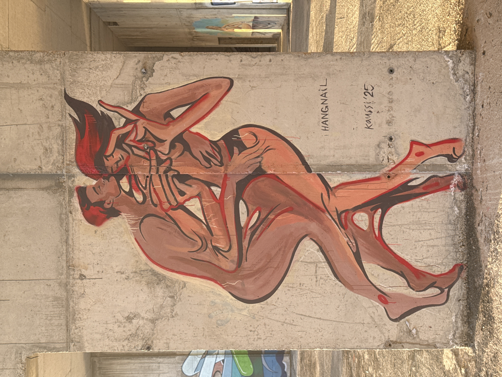 On a concrete wall in Perazića Do, Montenegro, is a mural depicting two figures intertwined in an embrace, painted in shades of reddish-brown and outlined in dark brown and red. The figures appear nude, with fluid, stylized forms. A name is visible above the mural, reading "HANGNAIL" with the signature “Kaussi ‘25” to the right. The concrete wall shows signs of weathering.