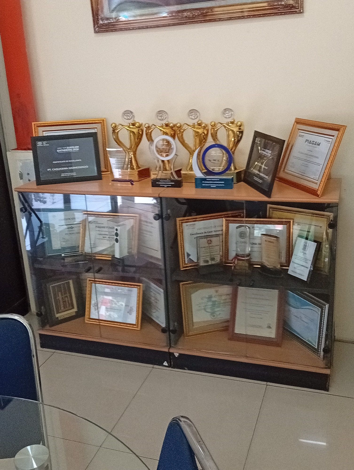The image shows a display cabinet with various awards and certificates, likely in an office or institutional setting. The cabinet is made of wood and glass, and it sits against a light-colored wall. On the top shelf of the cabinet, there are several golden trophies, some of which are decorated with clear glass elements. There are also framed certificates and a framed picture on this shelf.
The lower shelves of the cabinet are also filled with framed awards and certificates. These frames vary in color and material, including gold, wood, and possibly black. Some certificates are displayed upright, while others are tilted. Among the framed items, there are also some trophies and decorative awards. The cabinet has glass doors, which reflect the surrounding environment, including the floor tiles and what appears to be furniture in the foreground, such as a glass table and blue chairs. The overall impression is one of recognition and achievement being showcased. The location is specified as Harjamekar, Indonesia.