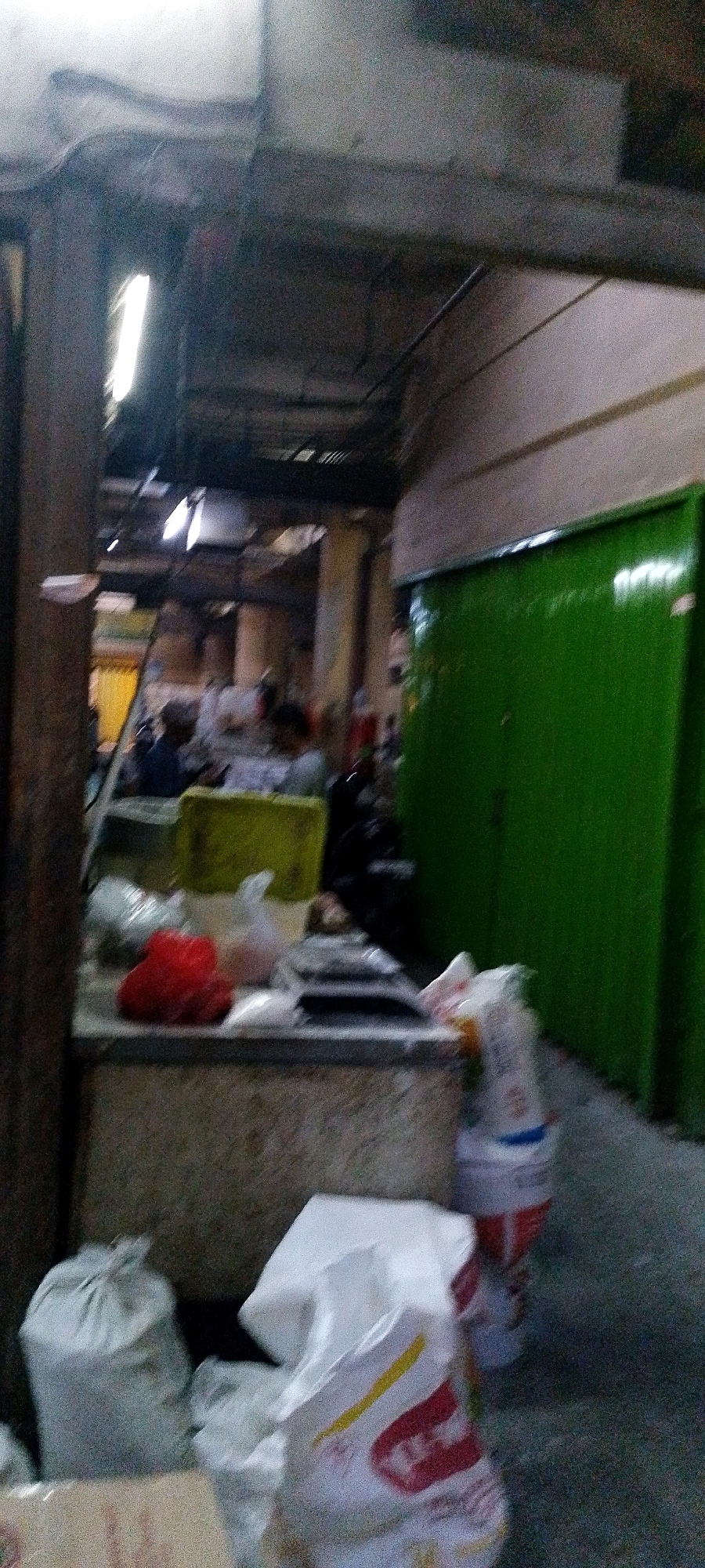 The image depicts a blurry indoor scene, likely a market stall in Bandung City, Indonesia. The environment appears to be dimly lit with overhead fluorescent lighting visible. In the foreground, a light-colored counter or table is laden with various items, including a bright red object, possibly fruit or vegetables, several plastic bags containing goods, and a dark rectangular object. Behind the counter, a large, vibrant green shutter or door dominates the right side of the frame, extending upwards and out of view. To the left of the counter, a wooden pillar is prominent, and further back, more stalls and people are visible in the background, though their features are indistinct due to the blurriness. Several sacks, one light gray and others white with red and yellow markings, are positioned on the floor in the lower part of the image.
