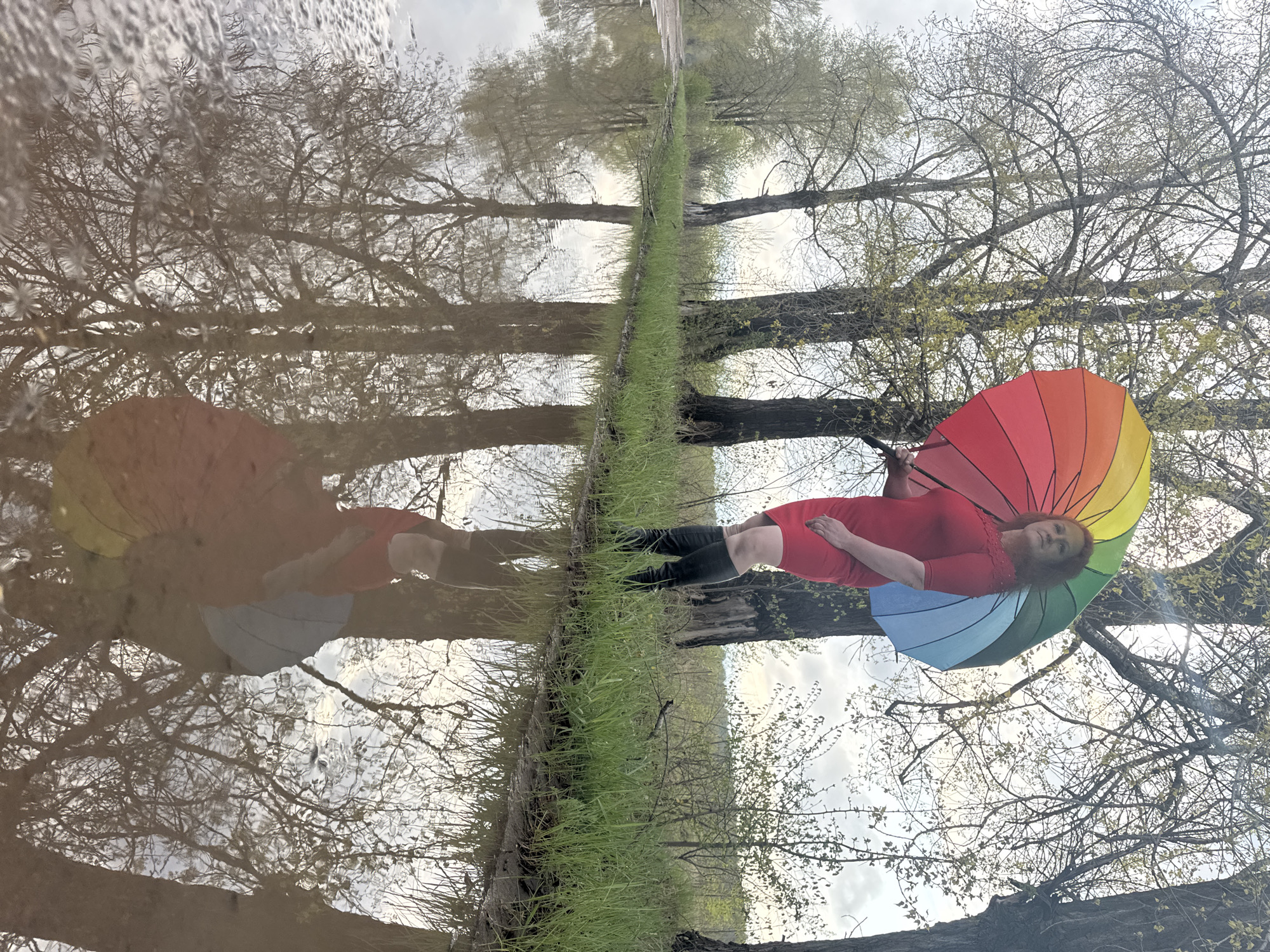 The image depicts a woman in Noviy, Russia, lying on a grassy bank next to a body of water. She is wearing a red dress and holding a rainbow-colored umbrella. The trees and sky are reflected in the water, creating a mirrored effect. The environment appears to be a park or natural area within the city. The woman is alone and appears to be posing for the image.