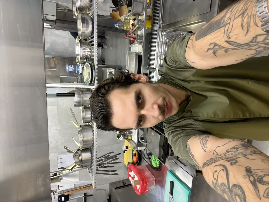 A male individual takes a selfie in a commercial kitchen environment. The subject, appearing to be in his 20s or 30s, has dark, wavy hair and a mustache, and is wearing a short-sleeved olive-green chef's jacket with dark buttons. Both forearms are extensively tattooed with black line art, including a snake, a skull, and other patterns. He looks directly at the camera with a neutral expression.
The kitchen features dominant stainless steel surfaces and equipment. On the counter to the left, a green cutting board with a knife, a red plastic container, a clear spray bottle, and a small dish of sliced red produce are visible. Centered in the mid-ground is a commercial panini press or grill. Above, a two-tier metal shelf unit holds numerous stainless steel pots, pans, and mixing bowls of various sizes. Beneath this shelf, multiple metal tongs hang from a rack. To the right, another section of stainless steel counter displays small metal containers with contents, oil bottles, and a metal grate with a basket. In the deeper background, a large blue water dispenser bottle is visible, and a blurry figure in dark trousers and light footwear is partially seen standing, indicating the presence of another person in the kitchen. The walls are composed of light grey tiles, and a large stainless steel exhaust hood is visible overhead. While the image depicts a commercial kitchen, there are no explicit visual identifiers within the photograph to confirm its location in Budva, Montenegro.