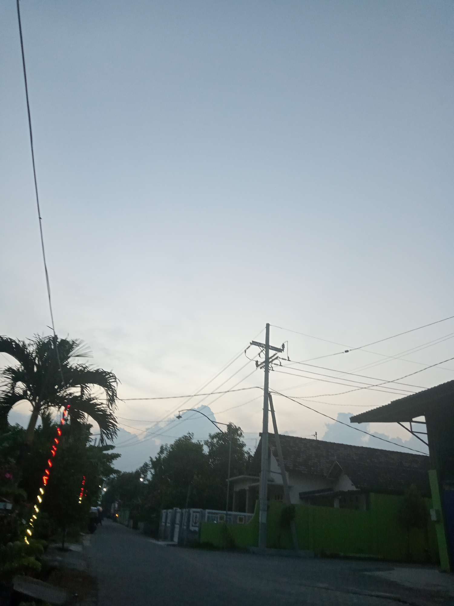 A low-angle view shows a street scene in Bungur, Indonesia. A utility pole with wires dominates the right side of the frame, positioned in front of a house with a brown tile roof and a green fence. Palm trees line the left side of the street, one decorated with vertical strings of red and yellow lights. In the distance, a road leads into the background. The sky is overcast with faint hints of cloud formation. A single motorcycle is visible in the distance.