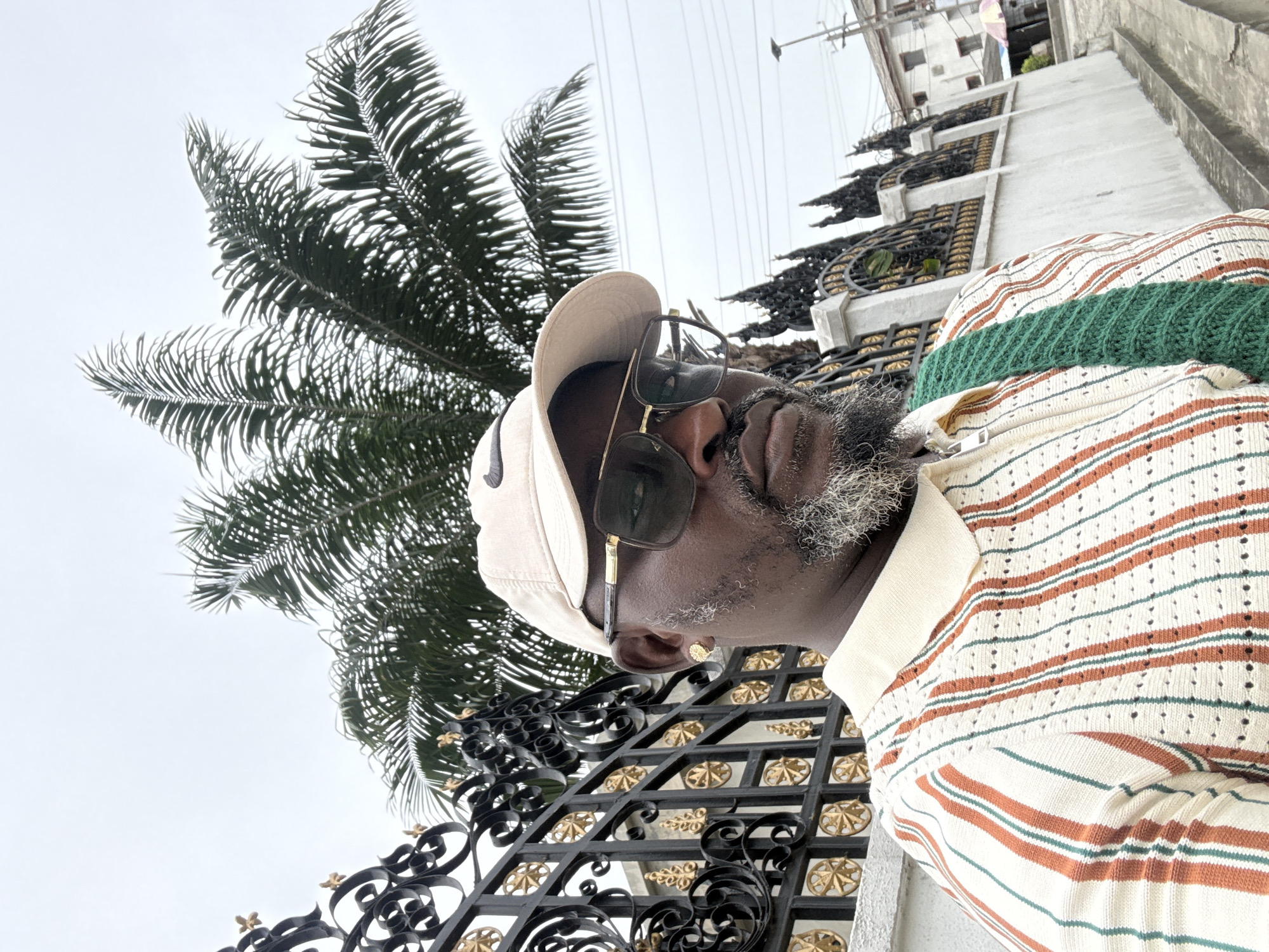 A medium shot shows a man from the chest up, wearing a tan baseball cap, sunglasses, and a striped shirt with a green knitted band across the chest. He has a gray beard and an earring in his left ear. The background includes a black wrought iron fence with gold accents, a palm tree, power lines, and a building. The location is Ado, Nigeria.