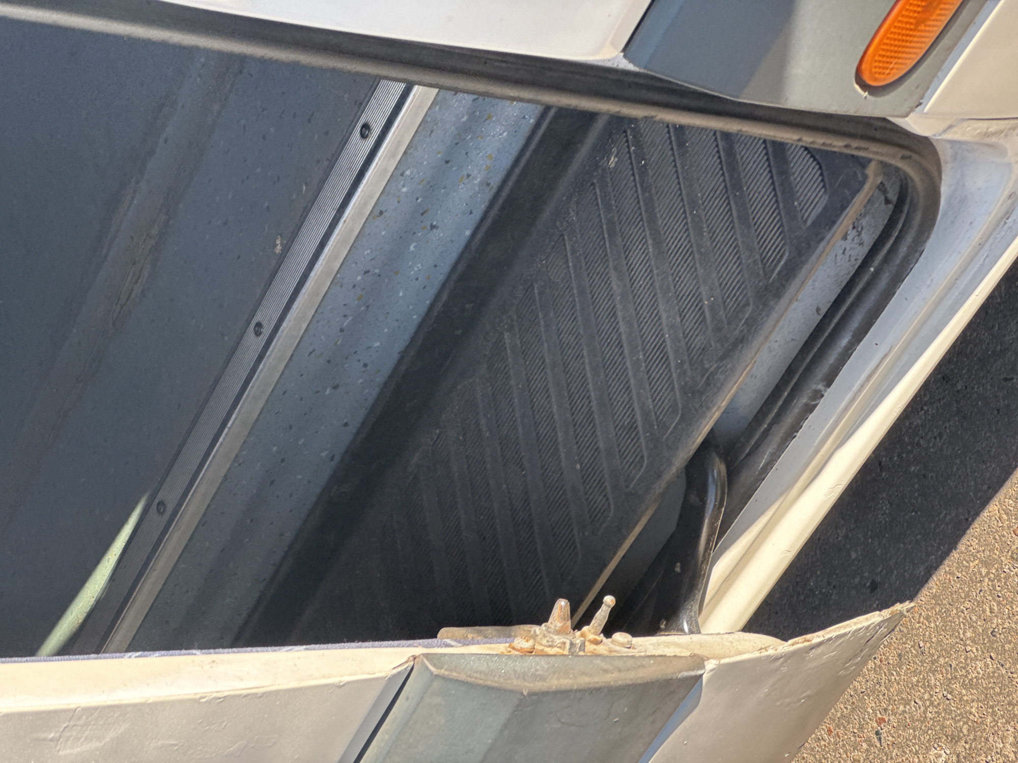 This is a close-up, eye-level, outdoor, daytime photograph of the interior step and threshold of a vehicle, likely a van or bus, with its door ajar. The step is covered with black rubber matting that has a ribbed texture. Above the rubber mat, there is a metal strip that appears to be part of the door frame or runner. The interior walls of the vehicle are a speckled blue-grey color. On the exterior of the vehicle, visible on the left side of the frame, is a weathered, light-colored door panel with rusted hinges. The ground outside the vehicle appears to be asphalt or gravel. A small, orange, reflective marker is visible on the exterior of the vehicle's body on the right side of the frame. The scene is located in the city of Kryvyi Rih, Ukraine.