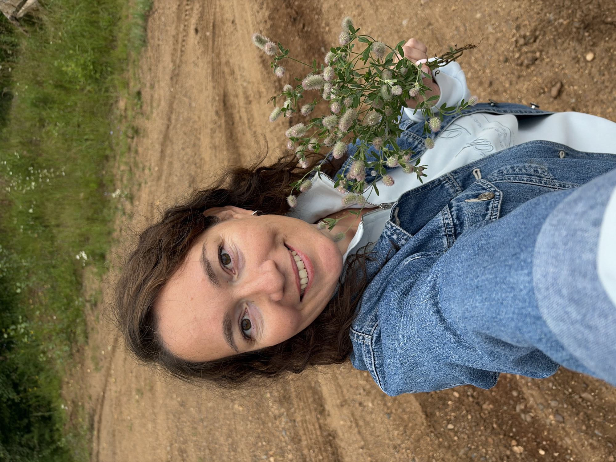 In an outdoor setting, a woman with dark, wavy hair and brown eyes smiles at the camera. She is wearing a denim jacket over a light blue sweatshirt. In her right hand, she holds a small bouquet of light pink and white wildflowers. The background consists of a dirt road and a grassy embankment with scattered white flowers. The image appears to have been taken in the city of Chernushka, Russia, given the context of the location.