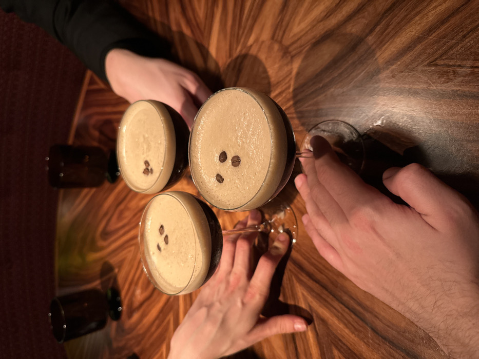 The image presents an overhead view of a social gathering around a warm-toned, polished wooden table with a distinct swirling grain pattern. Three hands are visible, belonging to at least two individuals, resting on or near the table. One hand on the right appears more masculine, while another at the bottom is more slender. A third hand, partially visible on the top left, wears a black sleeve.
The central subjects are three martini glasses, each filled with a creamy, light brown, frothy beverage, consistent with espresso martinis. Each drink is elegantly garnished with three dark coffee beans arranged on the foam. In the blurred background, two smaller, dark, empty glasses, possibly shot glasses or tumblers, are also visible.
The scene depicts an indoor setting, likely a bar, restaurant, or lounge in London, United Kingdom. The lighting is warm and somewhat dim, casting reflections and subtle shadows on the glossy table surface, suggesting the event is taking place in the evening or at night. The overall atmosphere appears relaxed and social, as individuals enjoy drinks together. There is no visible text in the image.