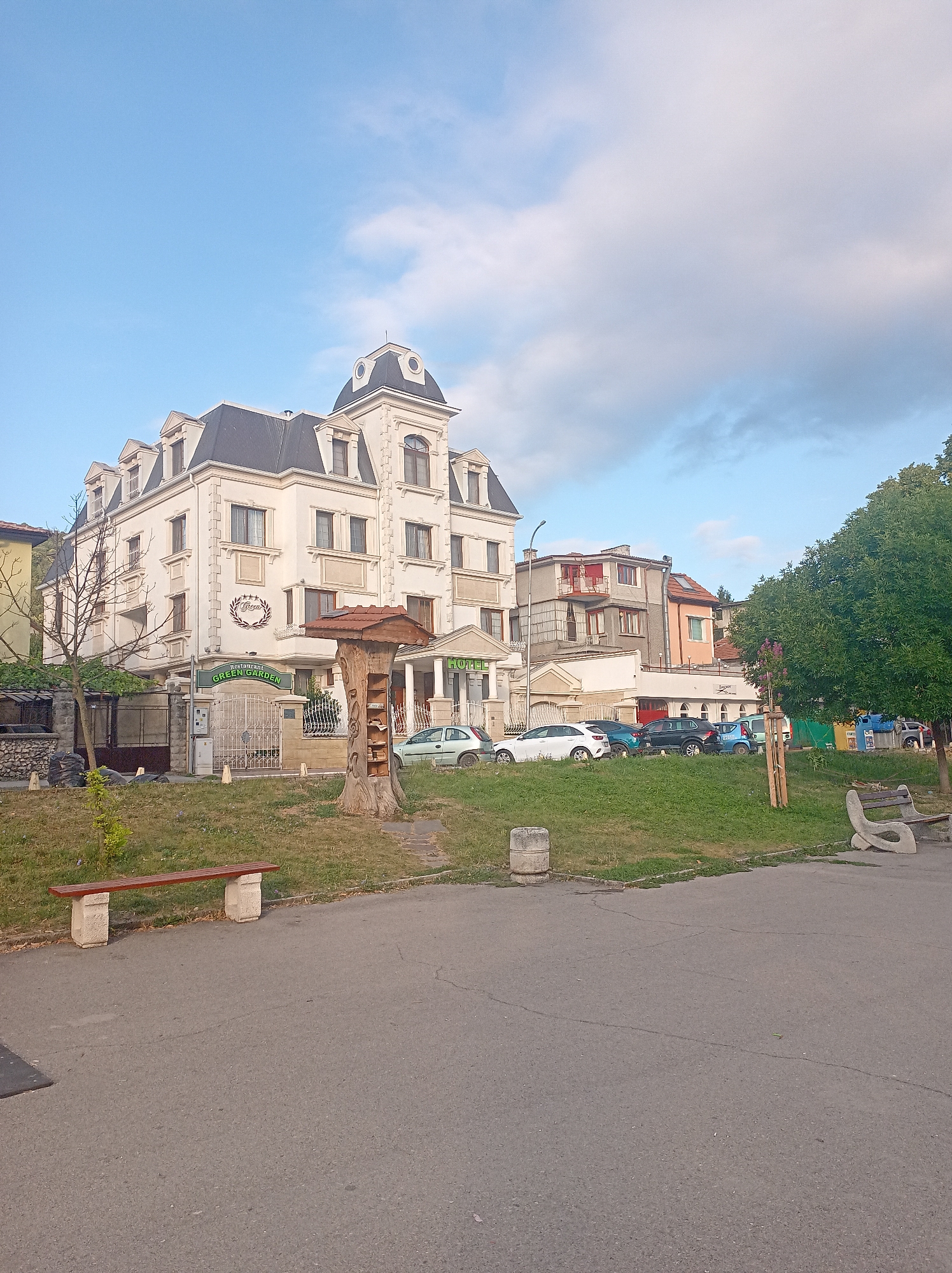 The image shows a street scene in a city. In the foreground, there is a paved walkway and a grassy area with a bench. A tree stump with a small book display is to the right of the building. The main building is a multi-story white structure with a distinctive mansard roof and decorative facade. A sign above the entrance reads "Restaurant GREEN GARDEN". Several cars are parked in front of the building and along the street. In the background, other buildings are visible, along with trees and a cloudy sky. The overall scene suggests a commercial area with a public space.