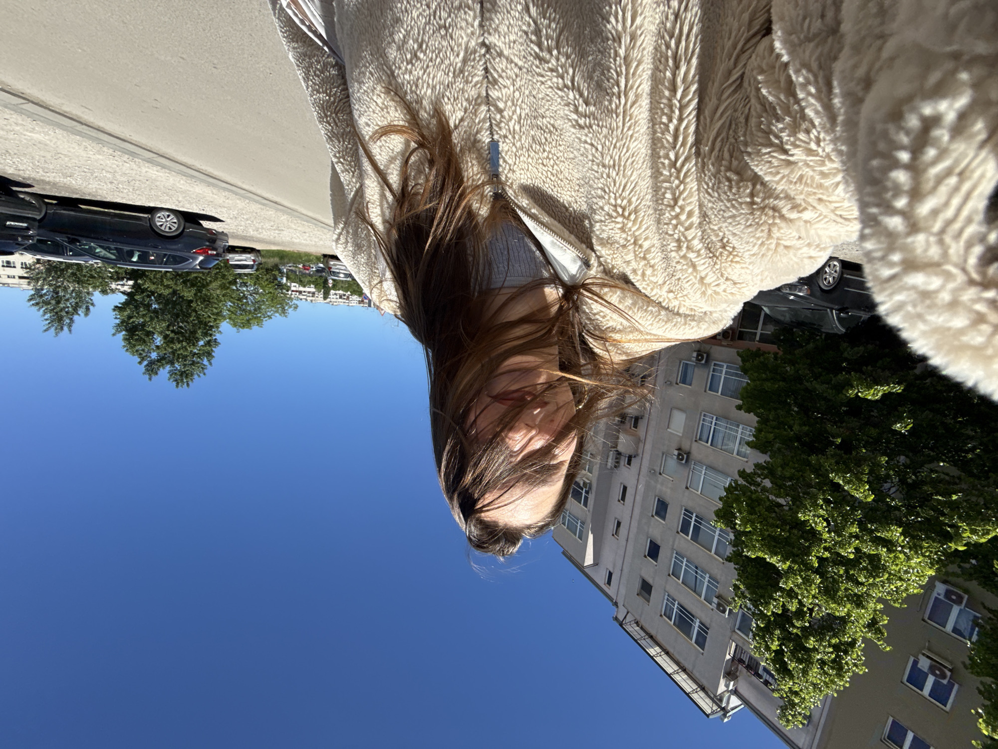 A young woman is standing on a street in Sofia, Bulgaria. She is wearing a fluffy, light-colored jacket and has long, brown hair. The sky is clear and blue. In the background, there is a tall, grey building with many windows and air conditioning units. A tree is partially obscuring the building. Across the street, there are parked cars and more trees. The photo is taken at an angle, making it appear as if the scene is tilted.