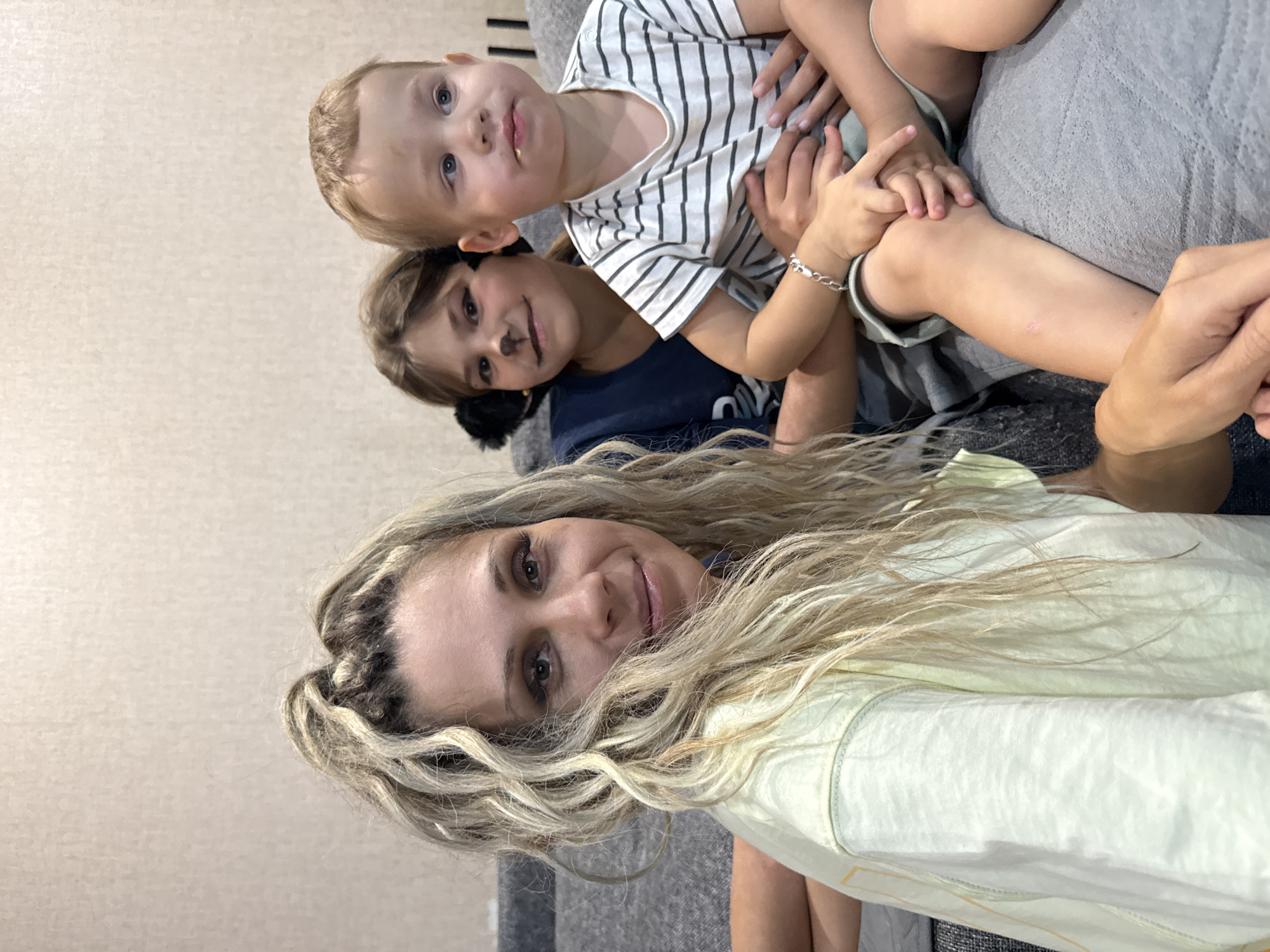 A woman with blonde, wavy hair is in the foreground, looking at the camera and smiling. She is wearing a light yellow t-shirt. Behind her, two children are seated. The child on the left has face paint resembling a cat's nose and whiskers, and is wearing a dark blue t-shirt. The child on the right is wearing a striped t-shirt and is looking to the right. The group appears to be sitting on a grey sofa. The background is a textured, light-colored wall. There are no discernible elements indicating a specific location in Rusanovo, Russia.