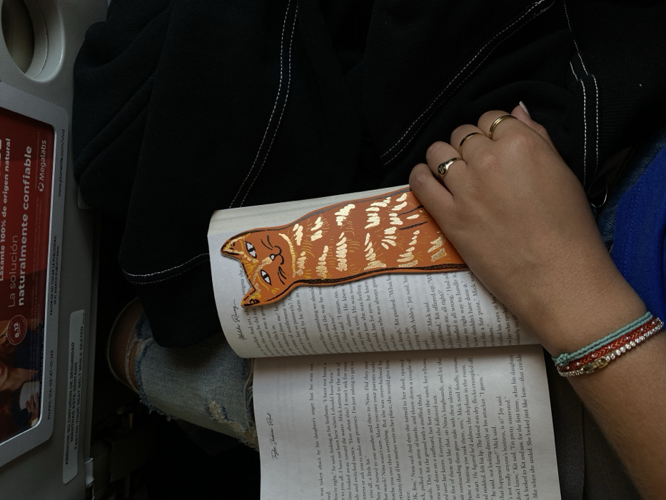 The image captures a top-down, angled view of a person reading a book, likely while seated on an airplane. The main subject is a person's right hand and arm holding an open book. The hand wears two gold rings, and the wrist is adorned with multiple bracelets, including beaded turquoise, red braided, and silver/rhinestone bands.
The open book features a distinctive orange cat-shaped bookmark with gold striped details, positioned at the top of the page. The text on the pages is legible in places, mentioning character names such as Mick, Nina, Kit, Ashley, and Jay, and phrases like "daughter's anger" and "elephant in the room," indicating a narrative story.
The person is wearing blue jeans with some distressed areas and a black jacket or top is draped nearby, with visible white stitching. To the left, an airline seat back or tray table is visible, along with an informational card. This card contains bilingual text in Spanish and English: "ABROCHE SU CINTURON DE SEGURIDAD MIENTRAS ESTE SENTADO" and "FASTEN YOUR SEAT BELT WHILE SEATED," confirming an aviation setting. Below this, another advertisement card is visible, promoting a "Laxante 100% de origen natural" (100% natural laxative) with the brand "Megalabs" and a website "www.planetite.com/viva". The overall setting is indoors, with no clear indication of the time of day.