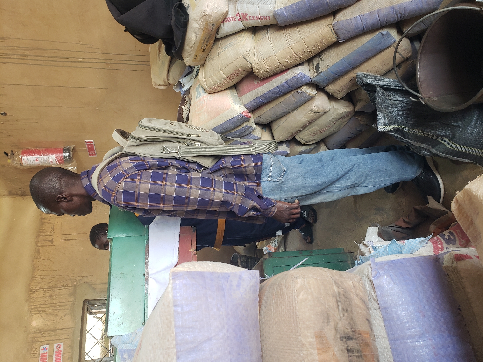 The image depicts an interior scene, likely a storage area or shop, with stacks of bagged goods, possibly cement or grain, filling the space. In the foreground, a man wearing a blue and brown plaid shirt and blue jeans stands facing to the right. He has a light-colored backpack slung over his left shoulder. Behind him, another person, appearing to be a child, is partially visible. The walls are a rough, textured, light brown. There are signs on the wall, one with red lettering indicating "NO SMOKING" and another with "ALCOHOL" and a red cross. A red fire extinguisher is mounted on the wall to the right of the "NO SMOKING" sign. A green counter or shelf runs horizontally across the middle of the image, with some items on top. Bags are stacked high on either side of the counter, some appearing to be wrapped in light purple material. On the floor in the foreground, a black bag and a metal bucket are visible. The overall impression is of a working or commercial environment. The location is identified as the city of Jos, Nigeria.