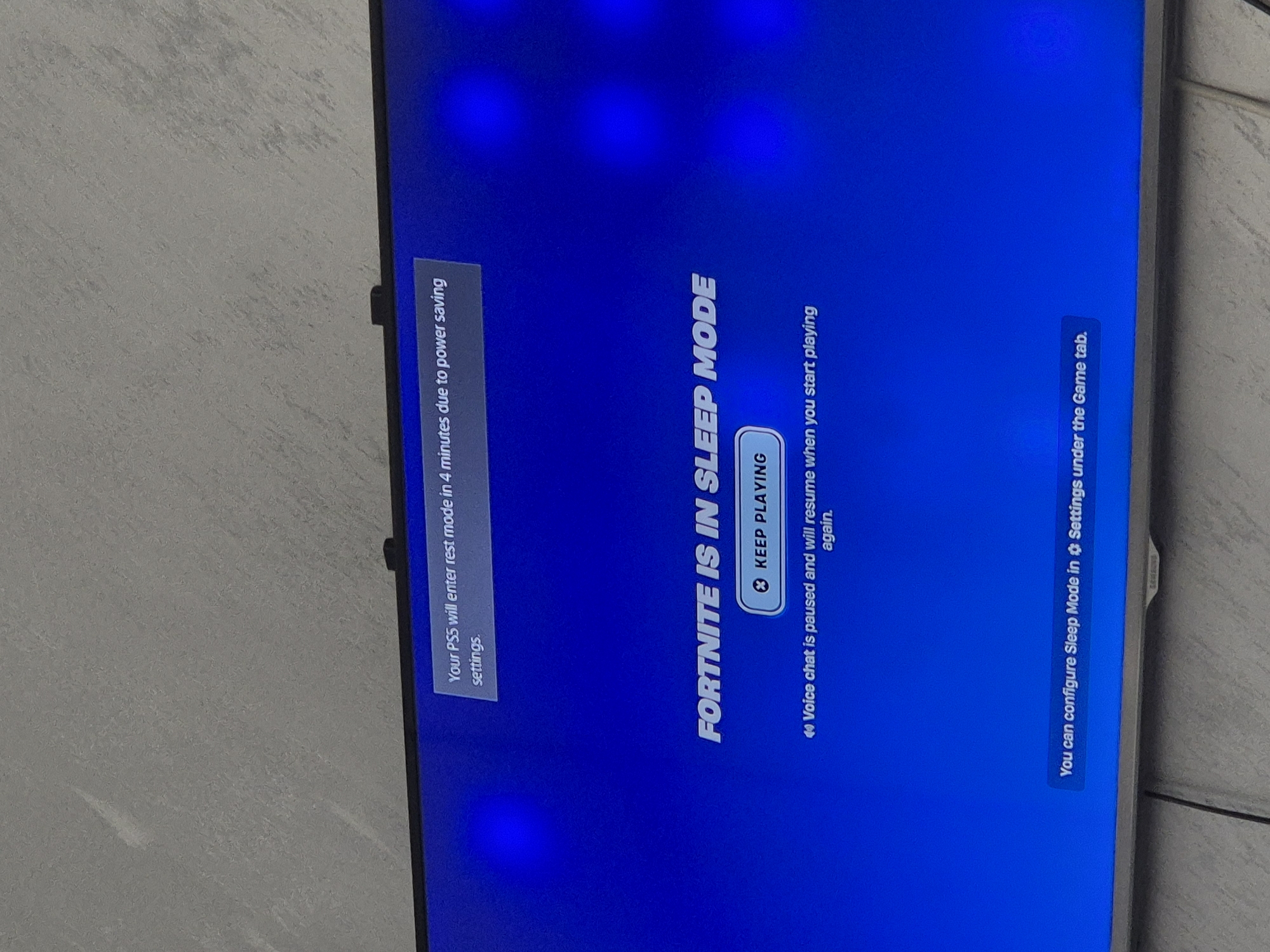 The image shows a television screen displaying a blue screen with white text indicating that "FORTNITE IS IN SLEEP MODE." A button labeled "KEEP PLAYING" is visible. Additional text states that voice chat is paused and will resume upon starting to play again, and that the sleep mode can be configured in the settings under the game tab. Another message informs the user that their PS5 will enter rest mode in 4 minutes due to power saving settings. The environment surrounding the television appears to be a simple, neutral-colored wall. No people or actions are present.