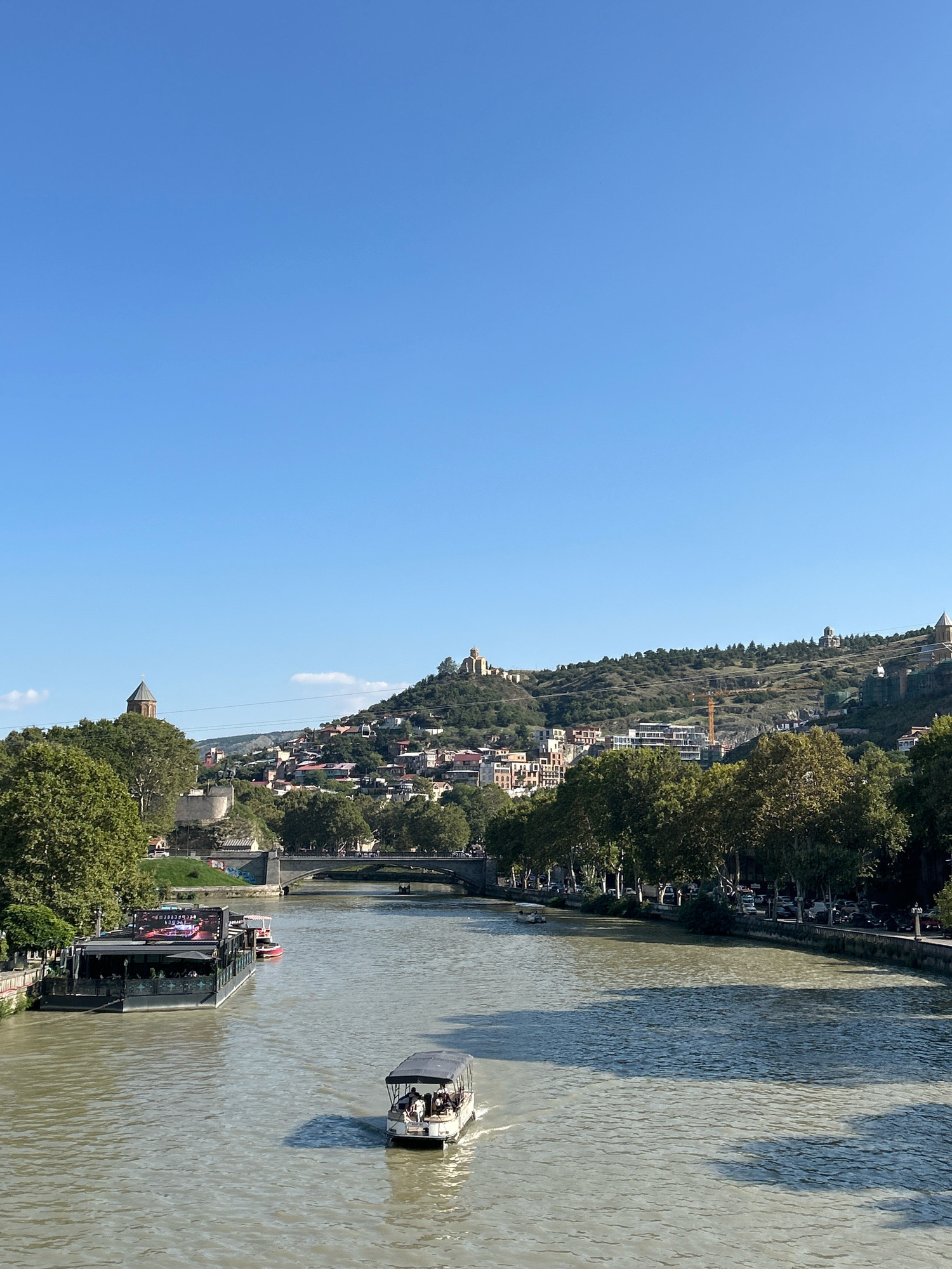 The image depicts a scenic view of the Mtkvari River in Tbilisi, Georgia, with the historic Old Town and Narikala Fortress visible on the hillside. The sky is a clear, bright blue, with a few faint clouds. The river is wide and the water is a murky greenish-brown color, reflecting the sunlight.
On the river, two boats are visible. The one closer to the foreground is a small, open-top tour boat with several passengers. It is moving away from the viewer and creating a wake. Further upriver, a larger, flat-bottomed barge is docked, which appears to be a floating restaurant or event space, with people visible on its deck. A red and white boat is tied to the barge.
Along the left bank of the river, trees with lush green foliage line the embankment. On the right bank, a street is visible with parked cars and more trees. The background is dominated by a steep, verdant hillside covered with buildings of varying architectural styles, indicative of the Old Town. Distinct structures on the hill include a church with a conical roof and a large fortress complex at the summit. A cable car line ascends the hill towards the fortress. The overall impression is one of a vibrant city situated amidst natural beauty and rich history.