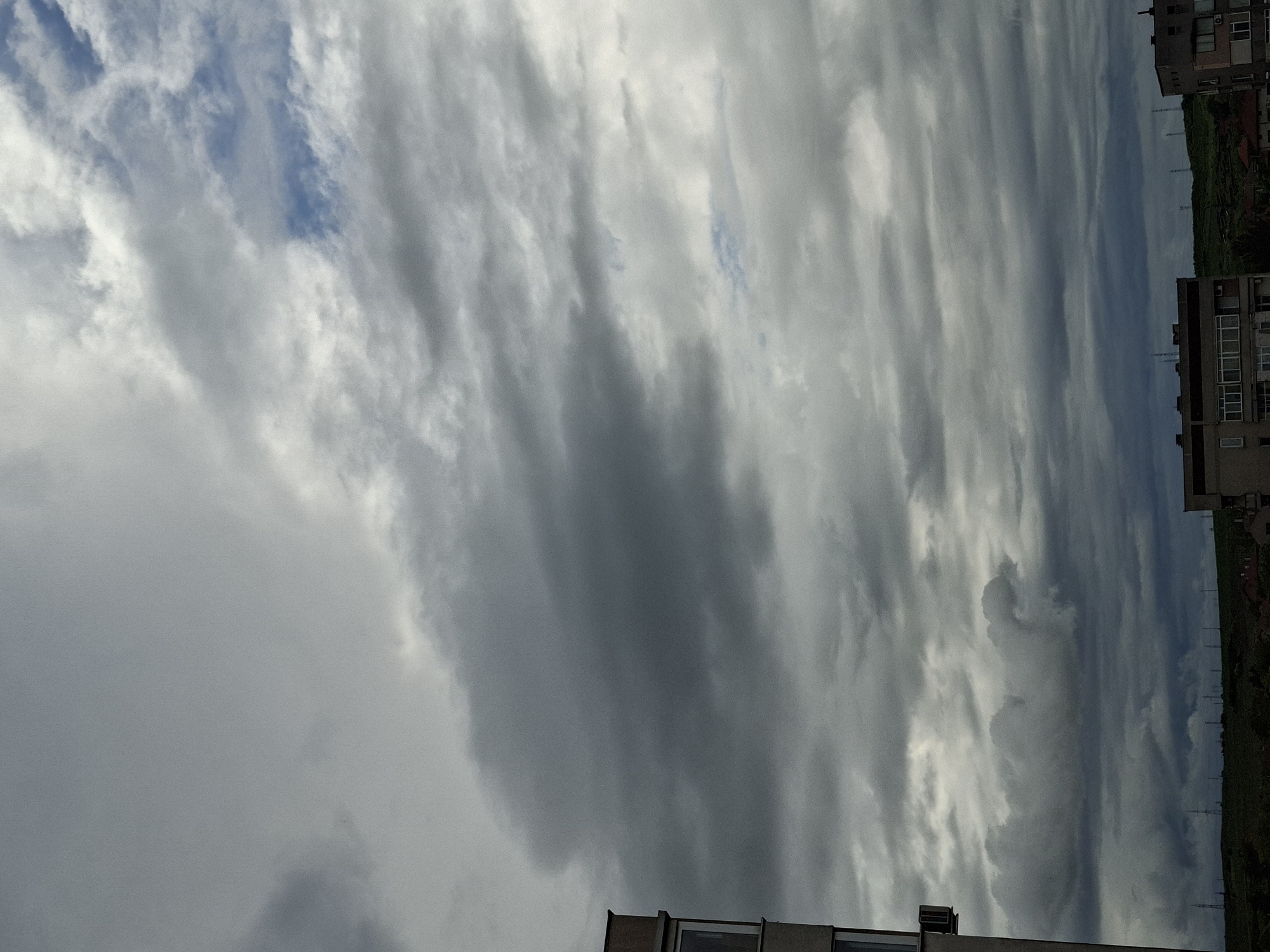 The photograph depicts a cloudy sky over Galabovo, Bulgaria. The sky is predominantly covered with gray and white striated clouds, with small patches of blue visible between them. A portion of a building is visible in the foreground at the bottom of the frame. On the right edge, buildings are present next to a green area with power lines. The orientation of the photograph is vertical.
