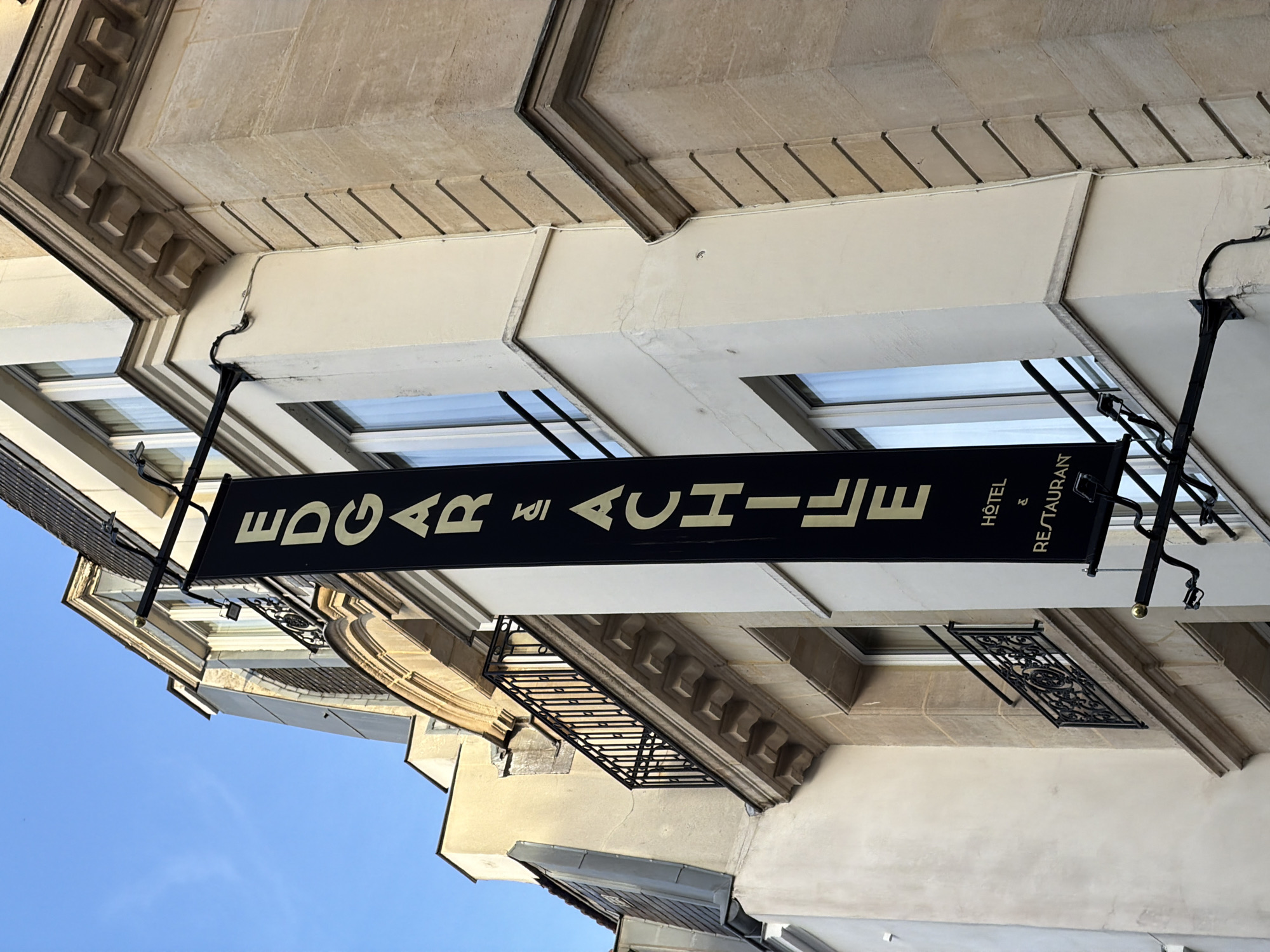The image shows a street-level view in Paris, France, looking upwards at a building facade. A black, rectangular sign hangs perpendicular to the building, displaying the name "EDGAR ACHILLE" in bold, sans-serif, gold-colored lettering. Beneath the name, in smaller print, it reads "HÔTEL & RESTAURANT." The sign is suspended by ornate black metal brackets attached to the building. The building itself is constructed of light-colored stone, with windows and decorative architectural details. A small balcony with a black wrought-iron railing is visible on one of the upper floors. The sky is a pale blue and is partially visible in the lower left corner of the frame.
