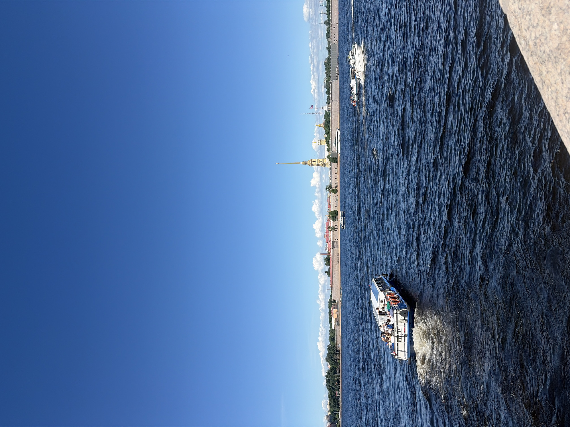 The image captures a wide outdoor scene on the Neva River in Saint Petersburg, Russia, on a clear, sunny day. The sky occupies the upper half of the frame, displaying a bright, cloudless blue with a few scattered white cumulus clouds along the horizon above the distant cityscape. The river's surface, filling the lower half, is dark blue and marked by numerous ripples and waves, indicating movement.
In the immediate foreground, a light-colored, possibly granite, embankment or wall is partially visible along the bottom right edge. A long, narrow passenger boat, featuring a white cabin and a blue hull, is positioned in the lower-left to mid-ground, moving away from the viewer towards the right. Several individuals are seated on the open top deck of this boat, and a white foamy wake trails behind it. Cyrillic text, possibly "МОЙКА", is visible on the boat's stern.
Further in the mid-ground, to the right of the passenger boat, a white motorboat with a visible wake travels horizontally across the river, heading right. A smaller boat is situated slightly behind and to its left. Near the distant riverbank, to the left of the prominent spire, a large white yacht is either moored or moving slowly.
The background features the distinctive architecture of Saint Petersburg, dominated by the Peter and Paul Fortress. A long, light-brown, fortified wall or embankment stretches horizontally across the scene. Rising prominently behind these fortifications, towards the left of the image, is the tall, slender, golden spire of the Peter and Paul Cathedral. Other buildings with red roofs are visible behind the fortress walls, interspersed with green trees. On the far right, further industrial structures, including a red-and-white striped chimney, are discernible on the distant bank. The entire scene is brightly illuminated by direct sunlight.