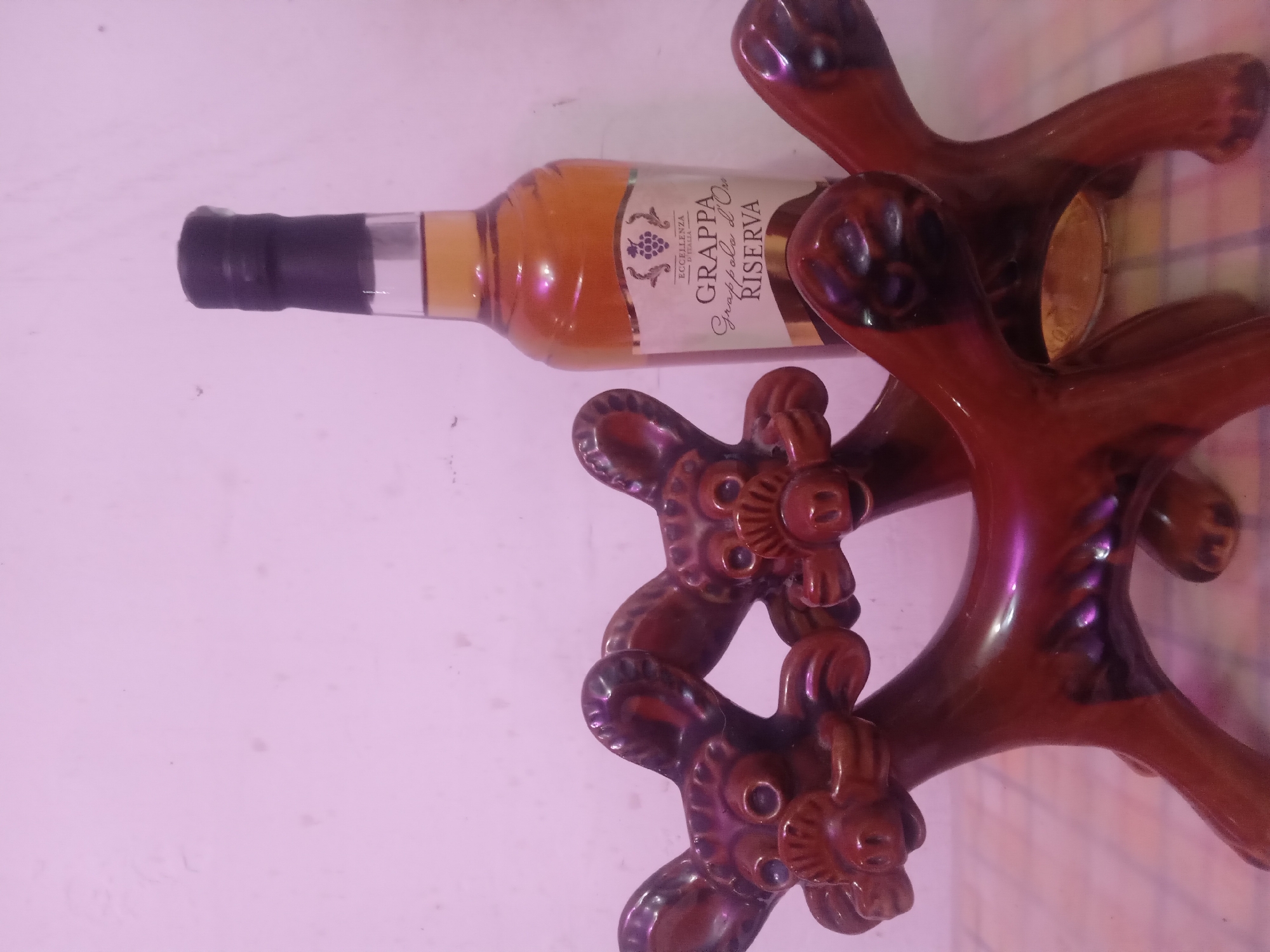 A still-life composition is captured under a pervasive pinkish-purple illumination, featuring a bottle of spirits and two decorative figures. The bottle, positioned in the mid-ground to the right, is transparent glass containing an amber liquid and sealed with a black cap. Its label is white and gold, displaying "ECCELLENZA D'ITALIA," a grape cluster graphic, and the text "GRAPPA Grappola d'Oro RISERVA." In the foreground, two dark reddish-brown, glossy ceramic figures are visible. These stylized, quadrupedal figures have prominent ears, circular eyes, and detailed snouts, and they partially obscure the lower section of the bottle. The objects rest on a light-colored surface with a faint grid pattern, which also reflects the dominant pink light. The background consists of a plain, pale pink wall with minor surface imperfections. The location in the city of Unknown, Unknown cannot be determined from the visual information provided.