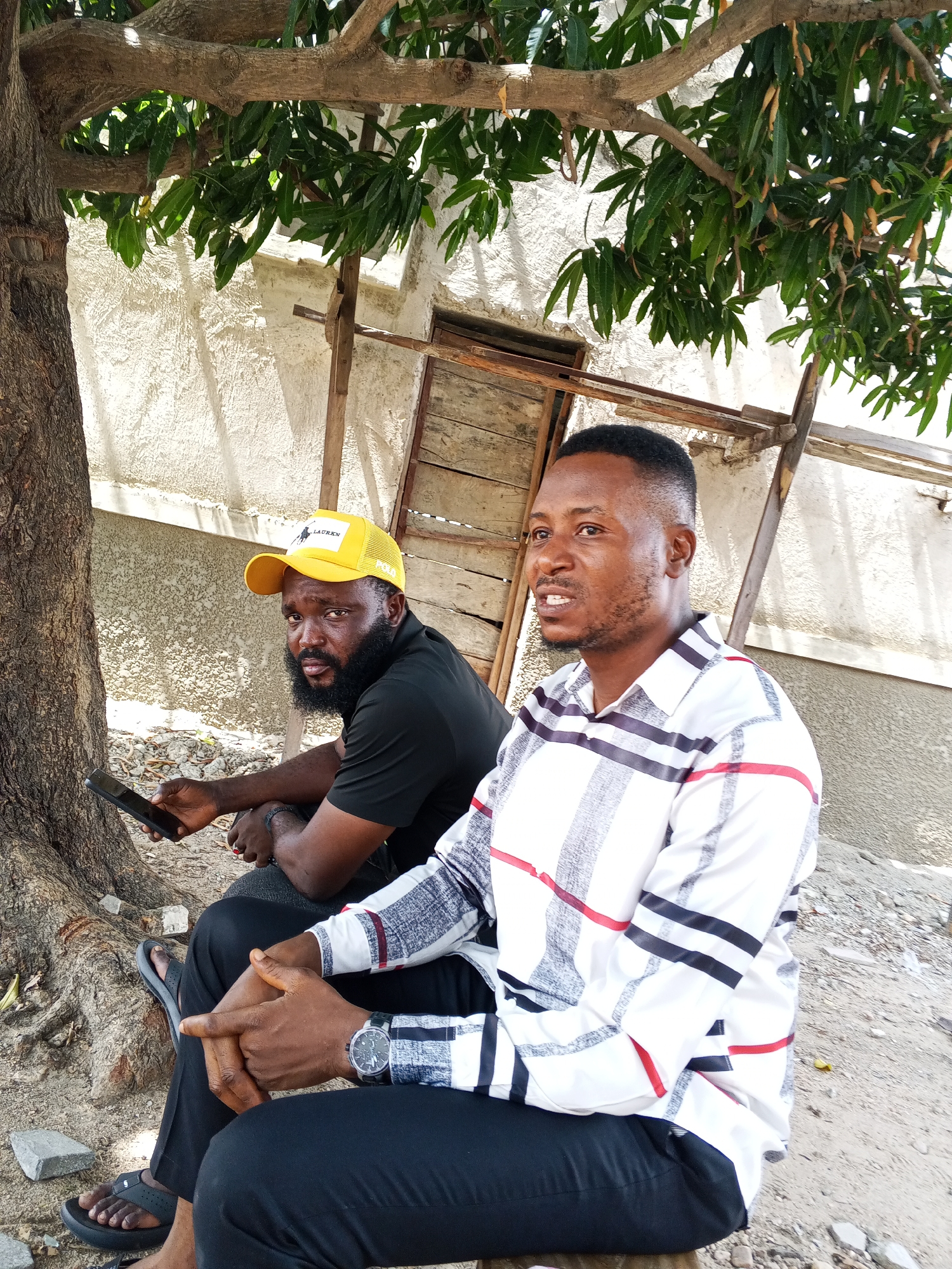 Two men are seated outdoors near a tree, possibly in Zango-ndaji, Nigeria. The man on the left wears a yellow baseball cap, a black t-shirt, and black sandals; he holds a mobile phone. The man on the right wears a plaid shirt, black pants, and a wristwatch; his hands are clasped. They are seated in front of a building with a boarded-up window and wooden scaffolding. The ground is partially covered with sand, small rocks, and tree roots. Branches and leaves overhang the scene.