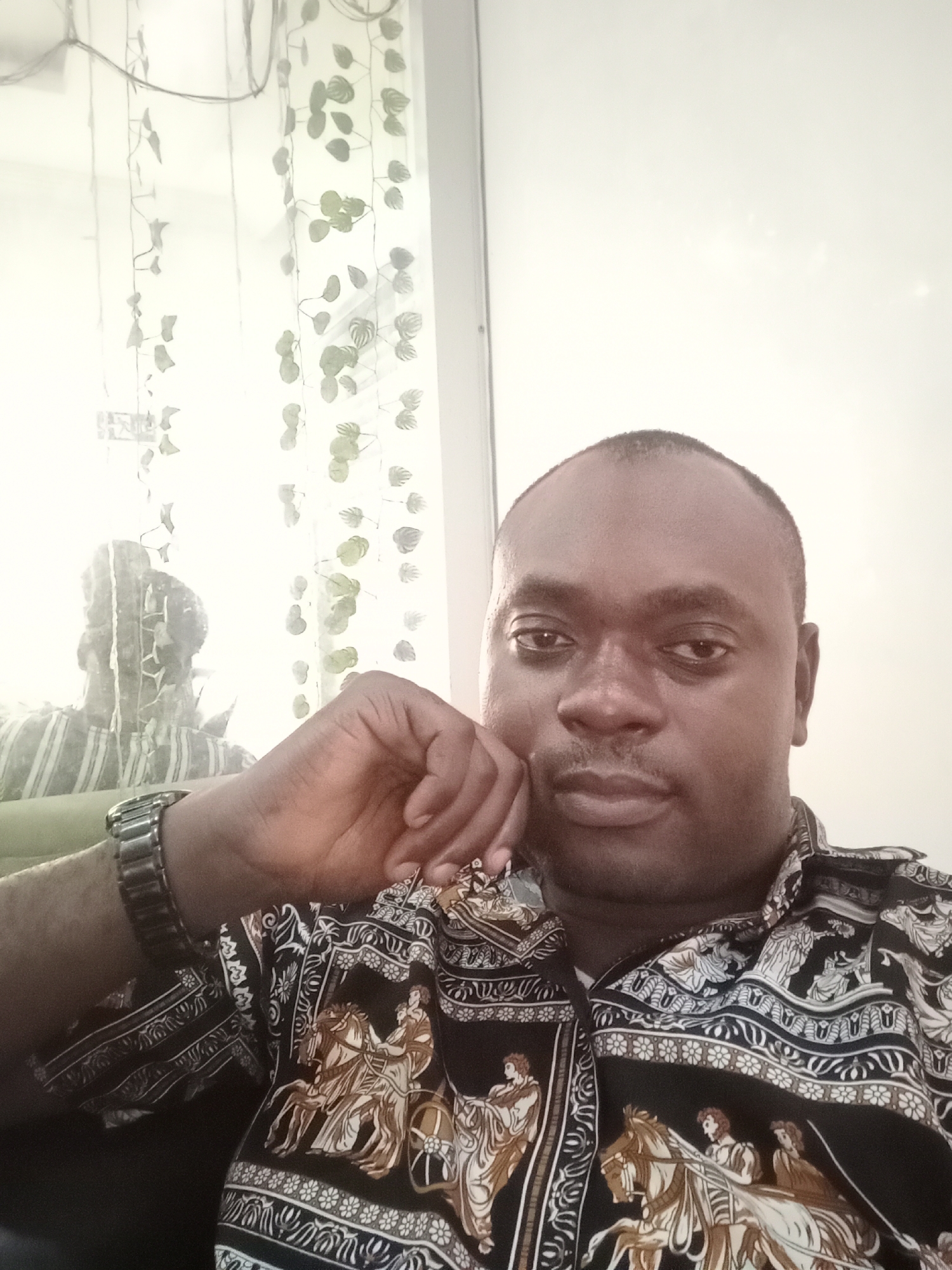 A man is captured in a medium close-up shot, appearing to be seated indoors. He has dark skin, a shaved head, and is wearing a patterned shirt featuring images of figures and horses, possibly depicting a chariot scene. A silver-tone wristwatch is visible on his left wrist as his hand is raised to his face. Behind him, there is a translucent curtain adorned with leaf-like designs. A faint reflection of another person in a striped shirt is visible in the curtain. The background is a plain, light-colored wall. The man's location is suggested to be in Gwarinpa, Nigeria.
