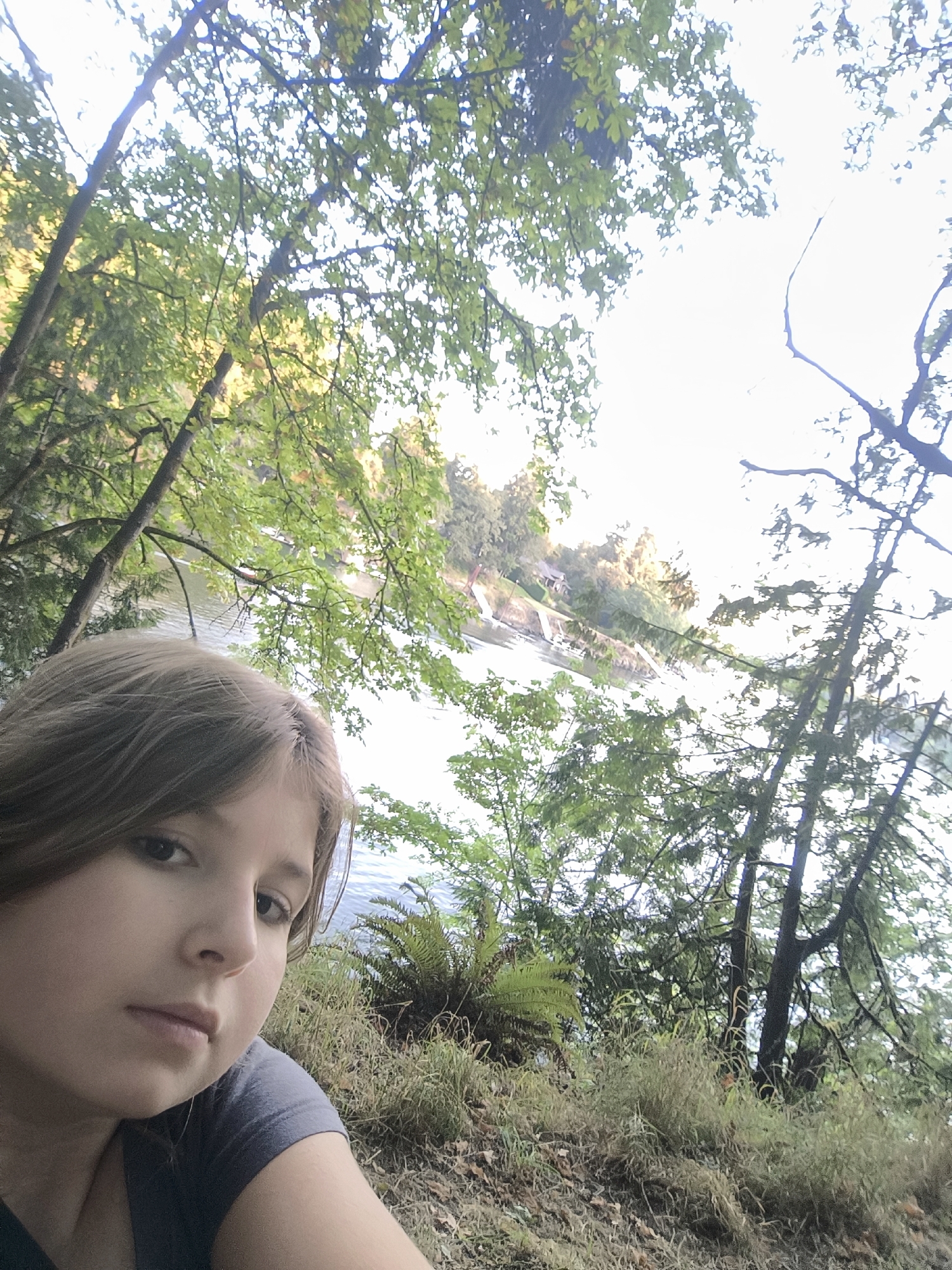 A young person with shoulder-length brown hair, wearing a grey t-shirt, is in the foreground of the image, looking slightly to their left. They are positioned on a grassy bank with dried leaves and ferns. The background features a body of water, likely a lake or river, with a tree-lined shore on the opposite side. Homes and a dock are visible on the far shore. The overall scene is outdoors, amidst a natural environment with lush green trees and dappled sunlight. The image appears to be taken in Lake Oswego, Oregon, United States of America.