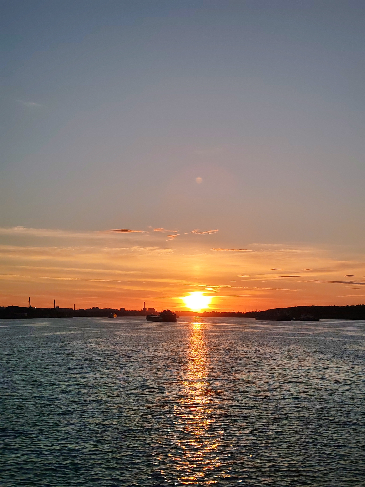 A scenic sunset paints the sky with vibrant hues of orange and yellow, casting a warm glow over a tranquil body of water. The sun, a brilliant orb, is setting on the horizon, its light reflecting on the water's surface, creating a shimmering pathway.
The scene is set by a vast expanse of water, with a silhouetted cityscape on the left and a forested shoreline on the right. Several ships are visible on the water, adding a sense of scale and activity to the serene landscape. One large cargo ship is prominently featured in the center, while other smaller vessels are scattered across the water. The sky above is a gradient of soft blues and oranges, with a few wispy clouds adding texture to the scene.
The overall mood is peaceful and serene, evoking a sense of calm and wonder. The time of day is clearly sunset, as indicated by the low position of the sun and the warm, golden light. The weather appears to be clear, with no strong winds or rain. The location is likely a coastal area or a large river, given the presence of ships and the extensive water body. There is no visible text in the image.