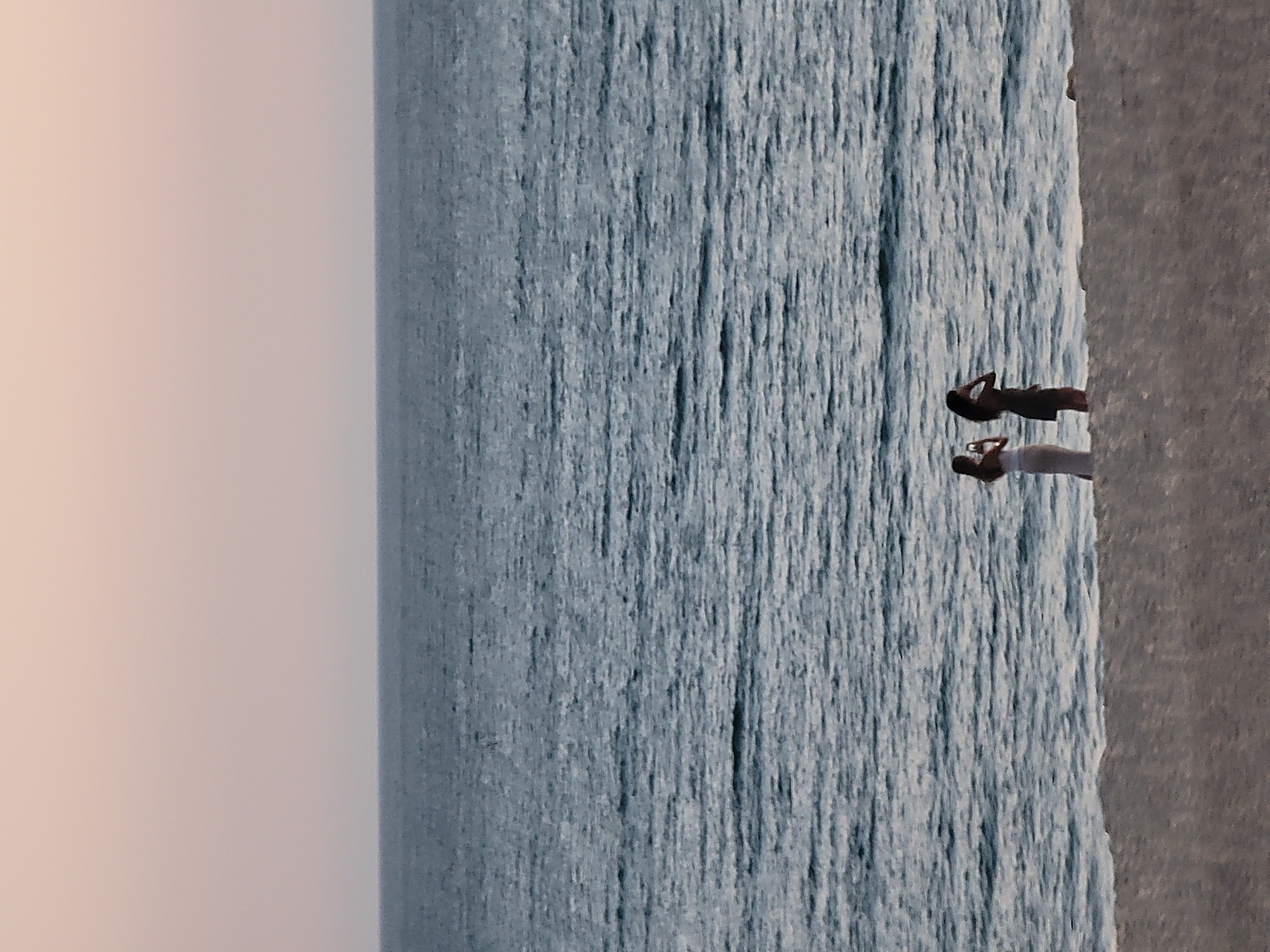 Two individuals stand on a sandy beach next to the ocean. The person on the left is wearing a white top and a light-colored skirt. They are holding a phone and appear to be taking a photograph. The person on the right is wearing a dark dress and has their hand near their head. The ocean is choppy with visible waves. The sky above is a soft, pale orange, suggesting either sunrise or sunset. The scene is captured from a slightly elevated perspective, looking down towards the water. The location is Quarteira, Portugal.