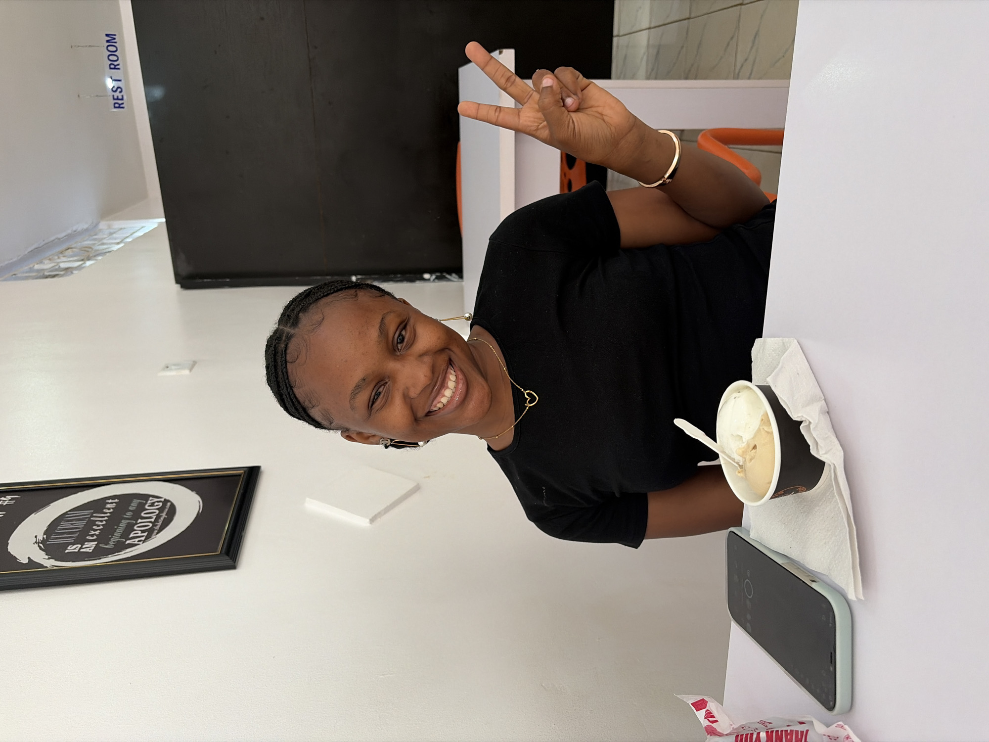 The image captures a cheerful young Black woman indoors, likely within a cafe or dessert shop in Bar Jirgi Summa, Nigeria. She is smiling broadly at the camera, with her head tilted, and her right hand raised to form a peace sign. She wears a black t-shirt, a gold bracelet on her right wrist, a gold necklace with a heart pendant, and small earrings. Her hair is styled in neat braids.
In her left hand, she holds a dark-colored cup of ice cream with a white spoon, which rests on a white counter or table. Next to the ice cream cup is a light blue smartphone, and a white tissue or napkin is also visible. The background consists of white walls and a large black panel. On the upper left wall, the words "REST ROOM" are clearly visible in blue lettering. Below the woman, on a white wall, there is a framed piece of wall art. Part of the text on the art reads: "AN EXCELLENT beginning to any APOLOGY." A fragment of a red and white plastic bag, partially displaying "THANK YOU," can be seen at the bottom right corner of the frame. The woman's expression conveys happiness and enjoyment, suggesting a relaxed moment.