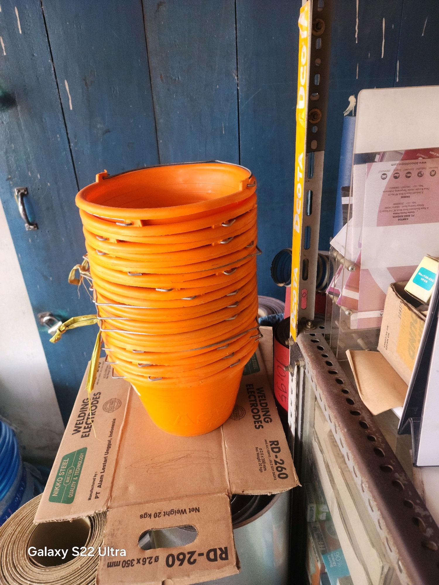 In an indoor setting with a weathered blue wooden door in the background, a stack of orange buckets is the primary focus. The buckets, arranged in a neat pile, are made of plastic and have silver wire handles. They are placed on top of a cardboard box. The cardboard box has text and graphics, including "WELDING ELECTRODES" and "NIKKO STEEL BUILT ON QUALITY & TECHNOLOGY." Further to the right, a metal shelf unit with visible rust and holes stands vertically. Various other items are visible in the background, including rolls of material and other boxes, suggesting a storage or workshop environment. The lighting is natural, casting shadows and highlighting the textures of the objects. The overall scene depicts a collection of industrial or utility items.