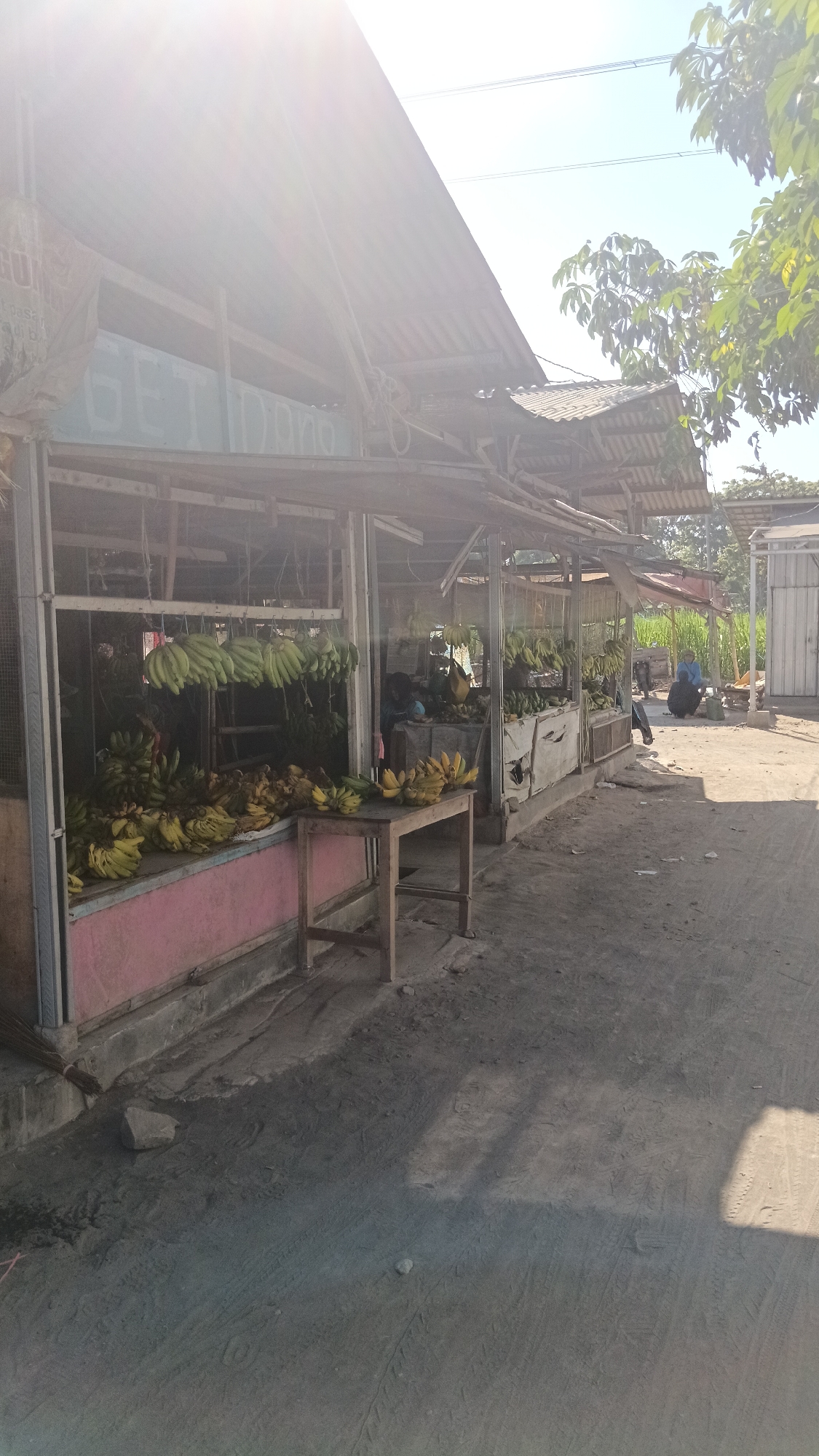 The image depicts an outdoor fruit stall located in Dukuh, Indonesia. The stall features a corrugated metal roof supported by wooden beams and metal framing. Various bunches of bananas are displayed, some hanging from the ceiling and others piled on tables and counters. A wooden table with bananas on it is placed in front of the stall. The ground in front of the stall is a dirt path with tire tracks visible. Two individuals are present at the stall, one appears to be a vendor or customer, and the other is seated further back. The environment suggests a sunny day with trees and vegetation visible in the background. The overall scene is characteristic of a local marketplace in a tropical region.