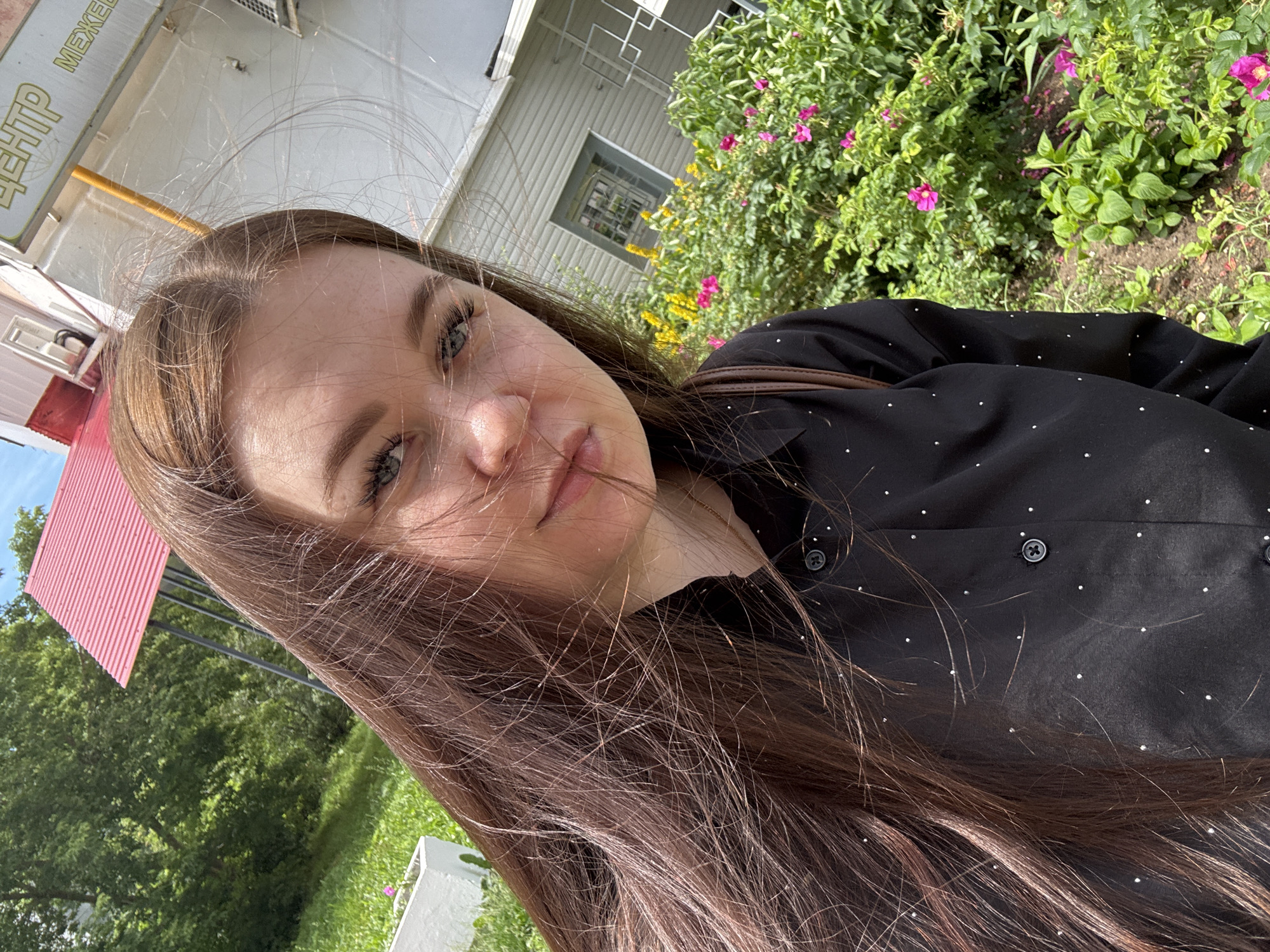 A young woman with long, brown hair and blue eyes is taking a selfie outdoors. She is wearing a black collared shirt with white polka dots. The background shows a building, a red roof, trees, and pink and yellow flowers. The image appears to be taken on a sunny day. The sign in the background, visible in the top right corner of the image, has text that is partially obscured but contains Russian lettering. Based on the visible text "ЦЕНТР" (Center) and "МЕЖЕ" (likely part of "Межевание" - land surveying, or "Межрегион" - interregional), and considering the architecture and vegetation, the location is likely a municipal building or a service center in Chaikovsky, Russia.