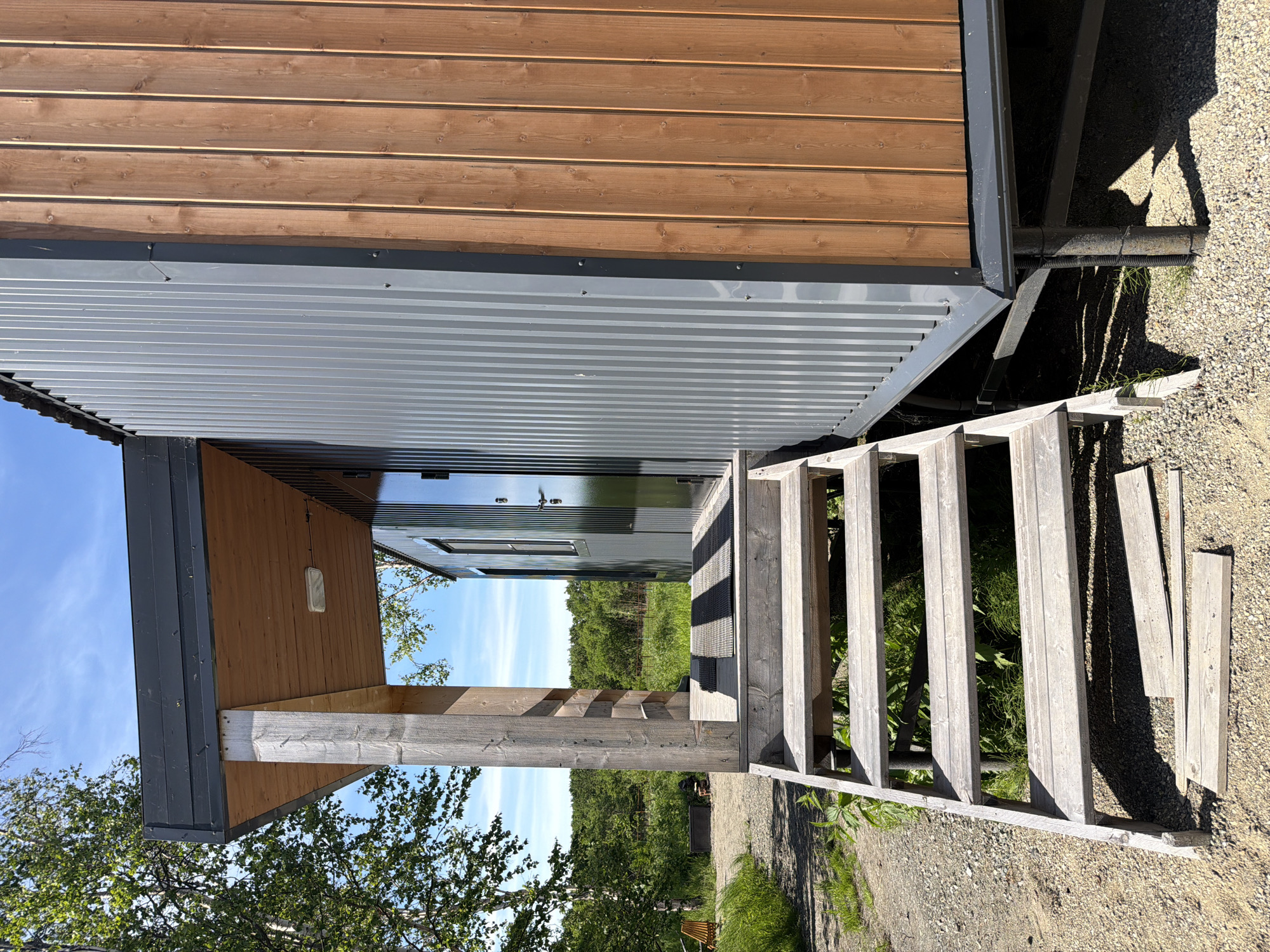 The image shows the exterior of a small, modern building with wooden steps leading up to a porch. The building is clad in corrugated gray metal and vertical wooden planks. A wooden support post holds up the covered porch. The area around the building is covered in gravel, with grass and trees visible in the background. The sky is blue and clear. There are no people or animals present in the image. The location is described as being in the city of Novy, Russia.