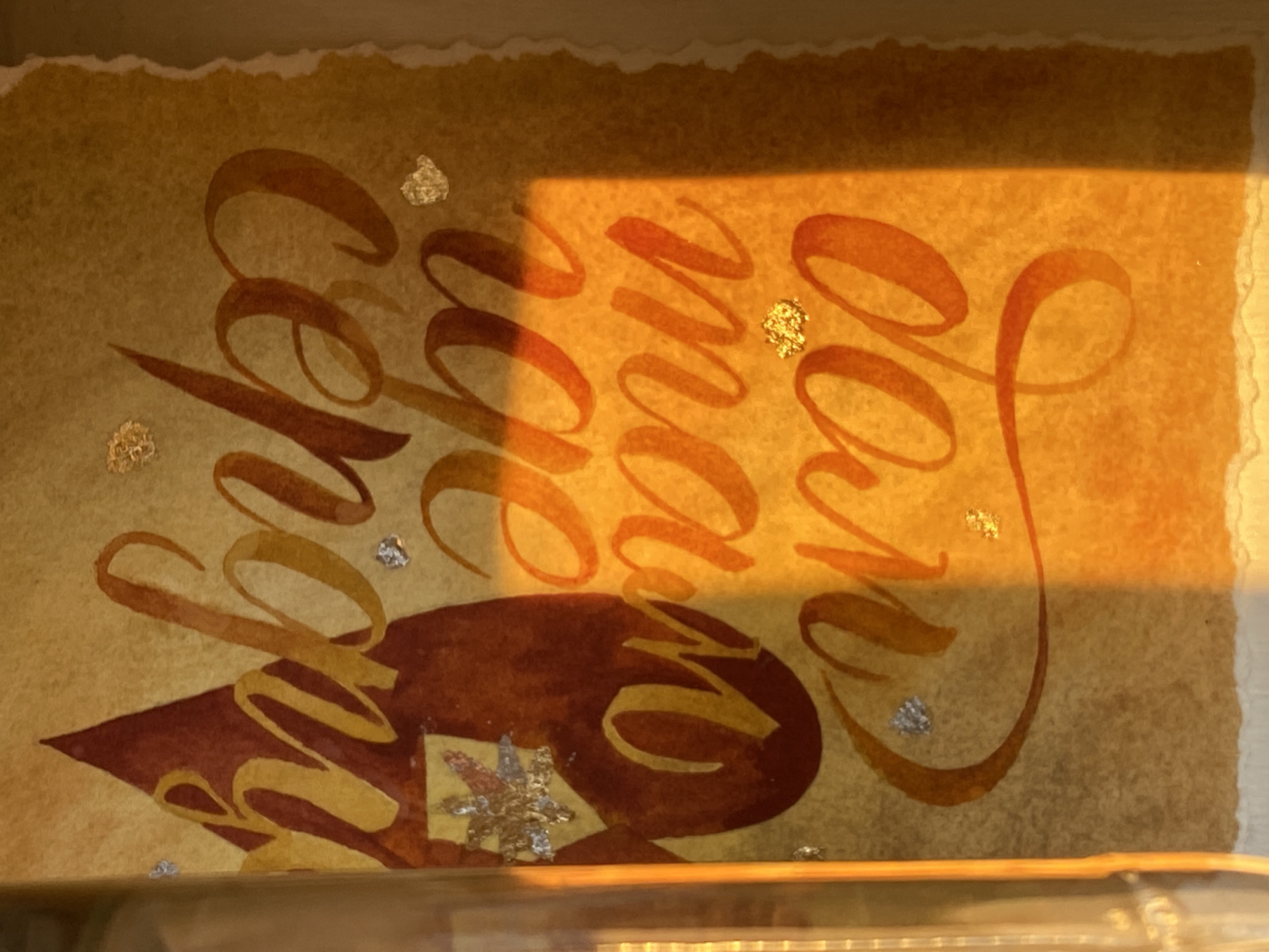 The image displays a calligraphic artwork rendered on a piece of textured, light brown or tan paper with rough, deckle edges. The paper is oriented vertically, and the calligraphy is written horizontally across it, appearing rotated 90 degrees counter-clockwise. A strong, golden sunlight casts a distinct, sharp shadow across the top and left portions of the artwork, indicating either early morning or late afternoon light.
The primary subjects are the paper and the intricate calligraphy. The text, rendered in shades of orange and reddish-brown, exhibits a gradient effect, with highlights of golden yellow, particularly where the sunlight catches the strokes. Interspersed among the words are small, irregular flakes of both gold and silver leaf, adding sparkle and texture.
The visible text, when mentally rotated to be read horizontally from left to right, appears to be the phrase "More You Love". The word "Love" is prominently placed on the right side of the composition.
Below the leftmost word ("More") and partially extending into the middle, there is a large, dark reddish-brown abstract shape, resembling a stylized mountain or a triangular form. Embedded within this shape is a decorative silver star.
The paper itself has a handmade, fibrous texture, visible in the illuminated areas. The bottom edge of the image shows a slight reflection, suggesting the artwork might be placed in a frame under glass or resting on a reflective surface. No specific activity or event is taking place; it is a static display of an art piece. The artwork is located in Chaikovsky, Russia.