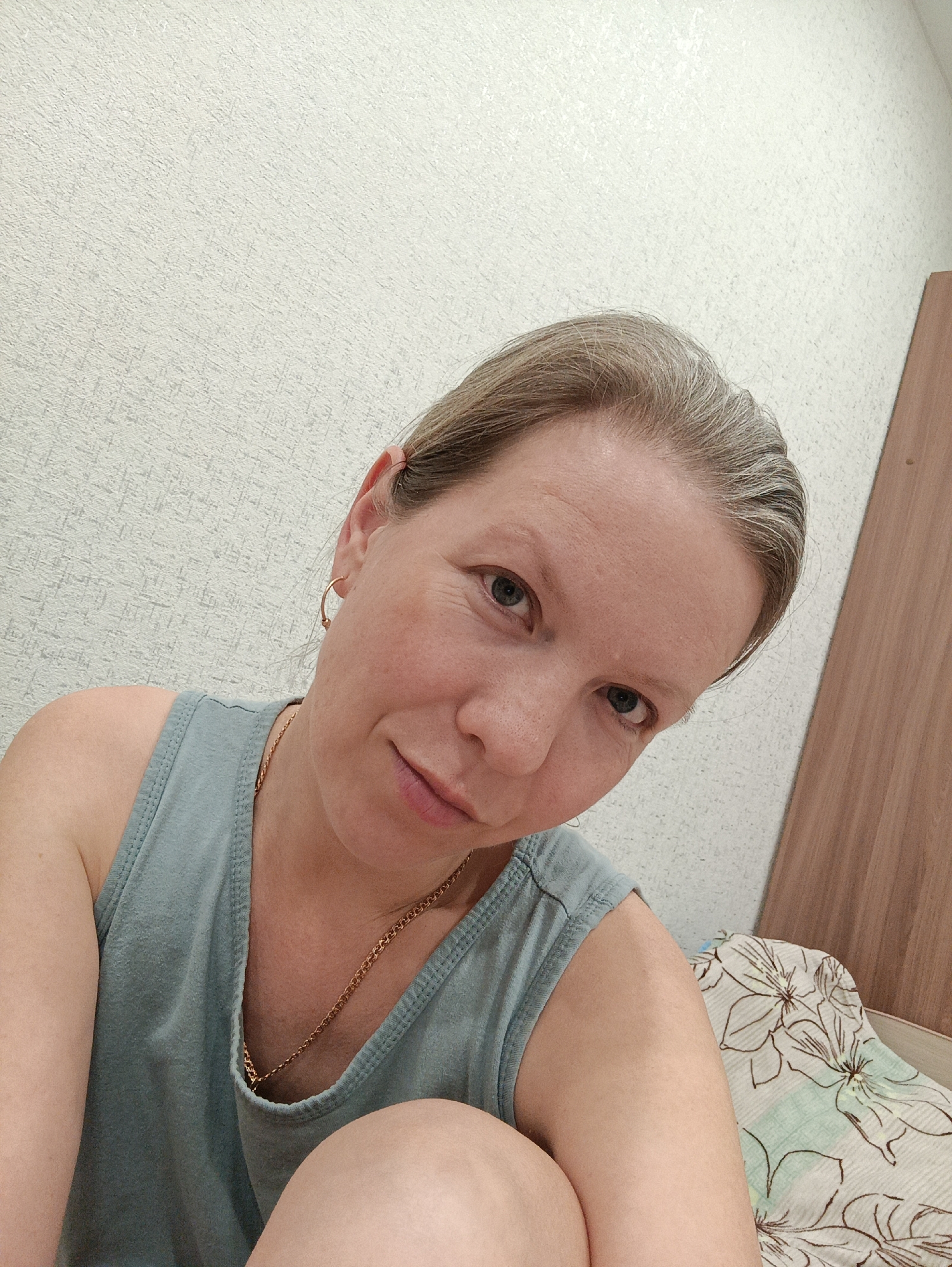 A woman with fair skin and light-colored hair is the primary subject of this photograph. She is wearing a blue tank top and a gold chain necklace. A small gold hoop earring is visible in her left ear. She appears to be sitting, with part of her arm and leg visible. The background includes a textured white wall and a section of a wooden cabinet or door. The woman is positioned in front of a bed covered with a patterned sheet featuring white and brown floral designs. There is no information in the image that indicates this photo was taken in Chaikovsky, Russia.