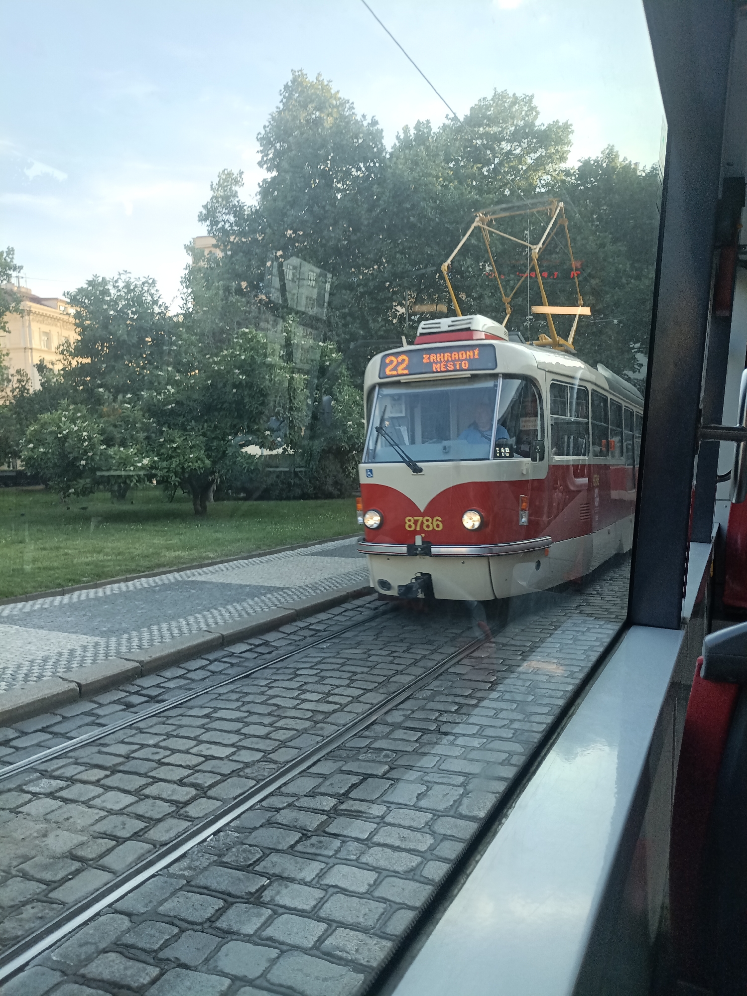 A red and cream colored tram with the number 22 and "ZAHRADNÍ MĚSTO" displayed on its front is traveling on a cobblestone track in Prague, Czechia. The tram has a driver visible through the windshield. The surrounding environment includes lush green trees and grass, with a paved walkway on the left side of the tram tracks. The image is taken from the perspective of someone inside another vehicle, likely another tram, given the reflection in the window. The sky is partly cloudy and light filters through the leaves of the trees.